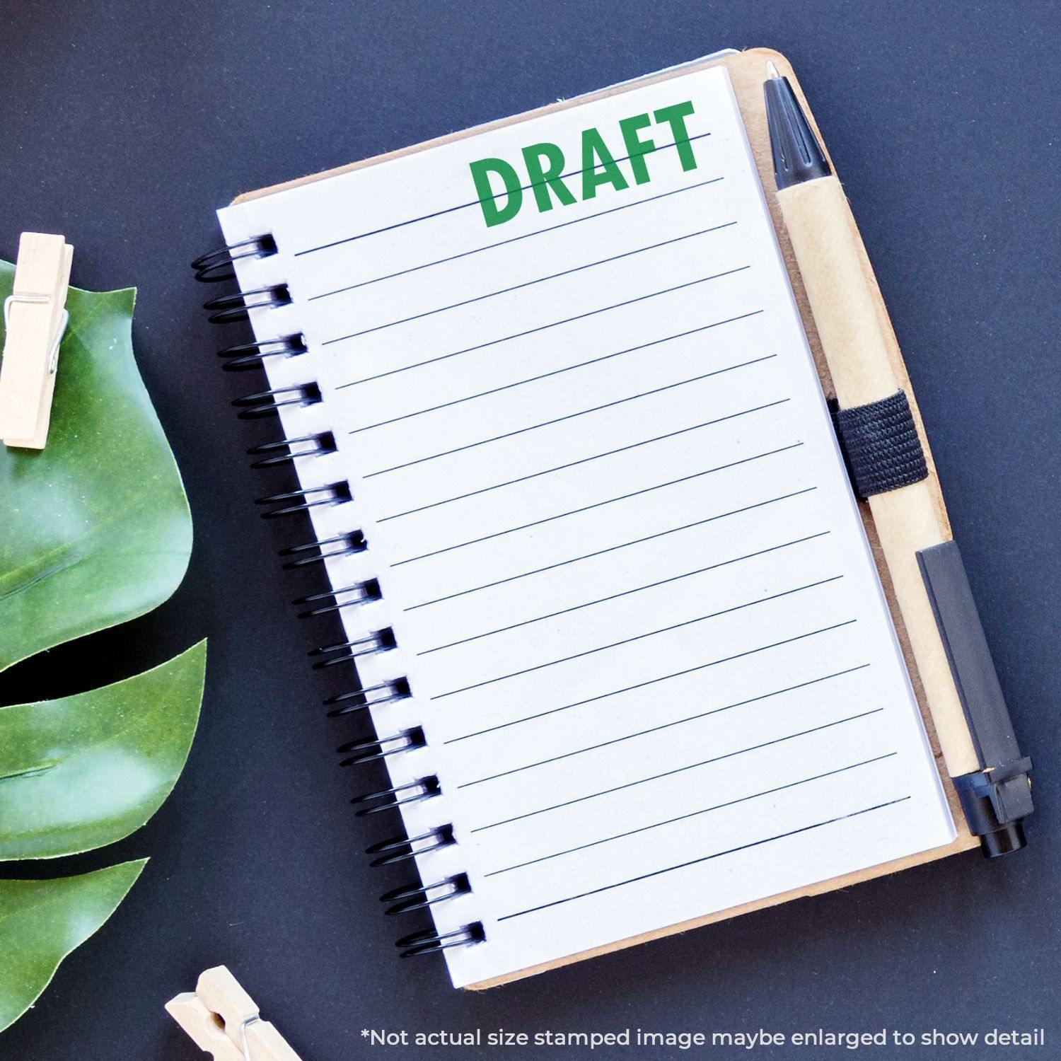 A notebook with a DRAFT mark from a Draft Rubber Stamp, placed on a black surface with a pen and green leaves nearby.