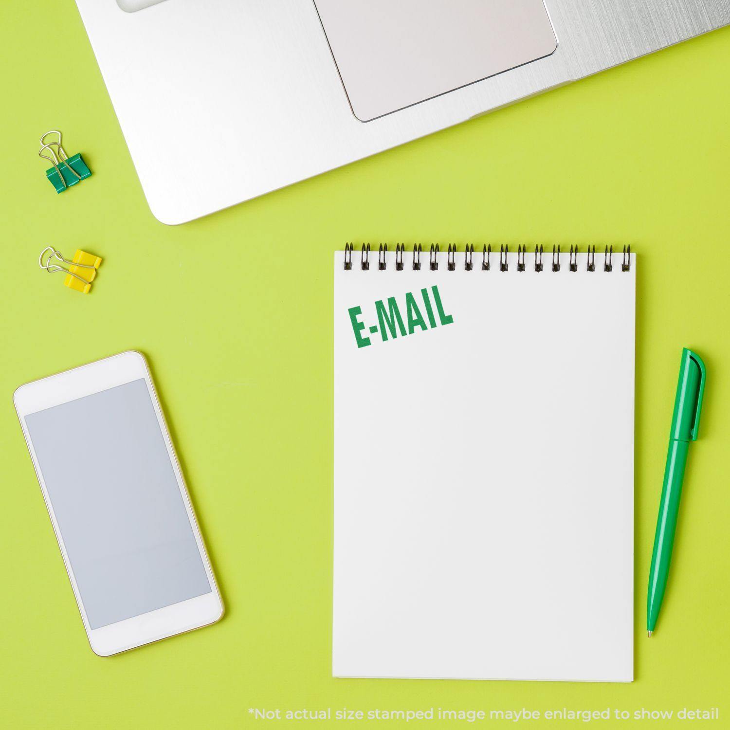 E Mail Rubber Stamp used on a white notepad next to a green pen, smartphone, laptop, and yellow binder clips on a green surface.