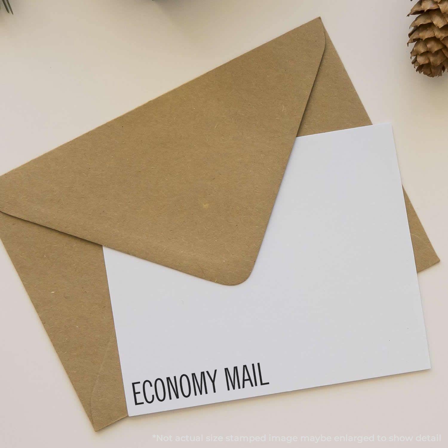 Large Pre-Inked Economy Mail Stamp used on a white envelope, placed on a beige surface with a brown envelope and a pinecone nearby.