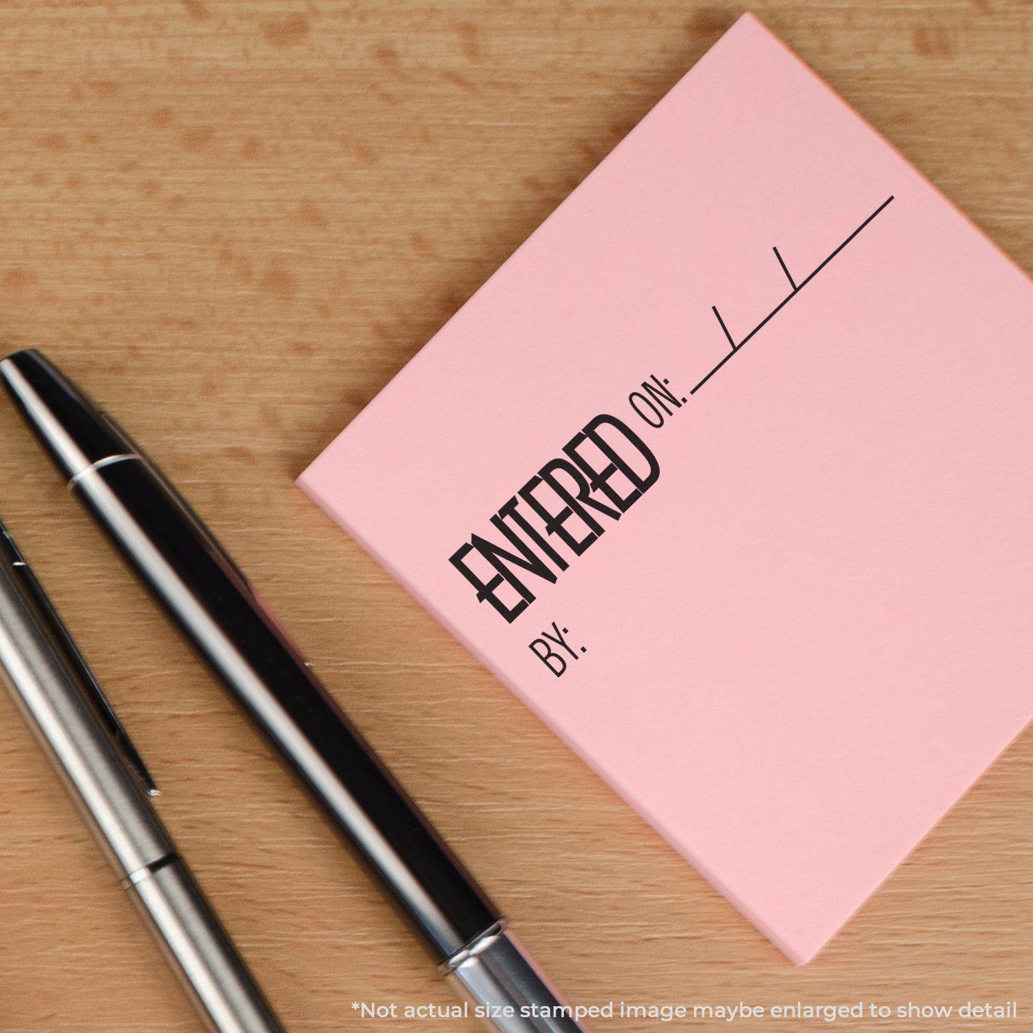 Large Self Inking Entered On Stamp on a pink sticky note, next to two pens on a wooden surface.