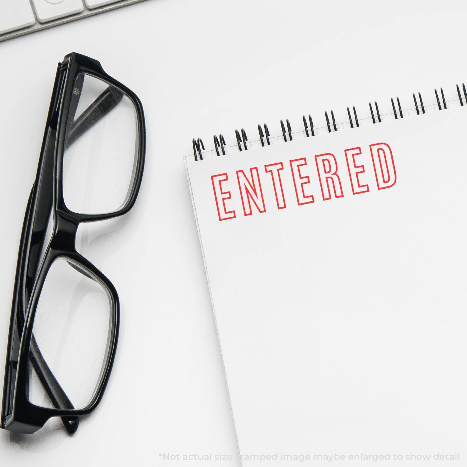 Large Self Inking Entered Outline Stamp marking 'ENTERED' in red on a spiral notebook, with glasses and a keyboard nearby.