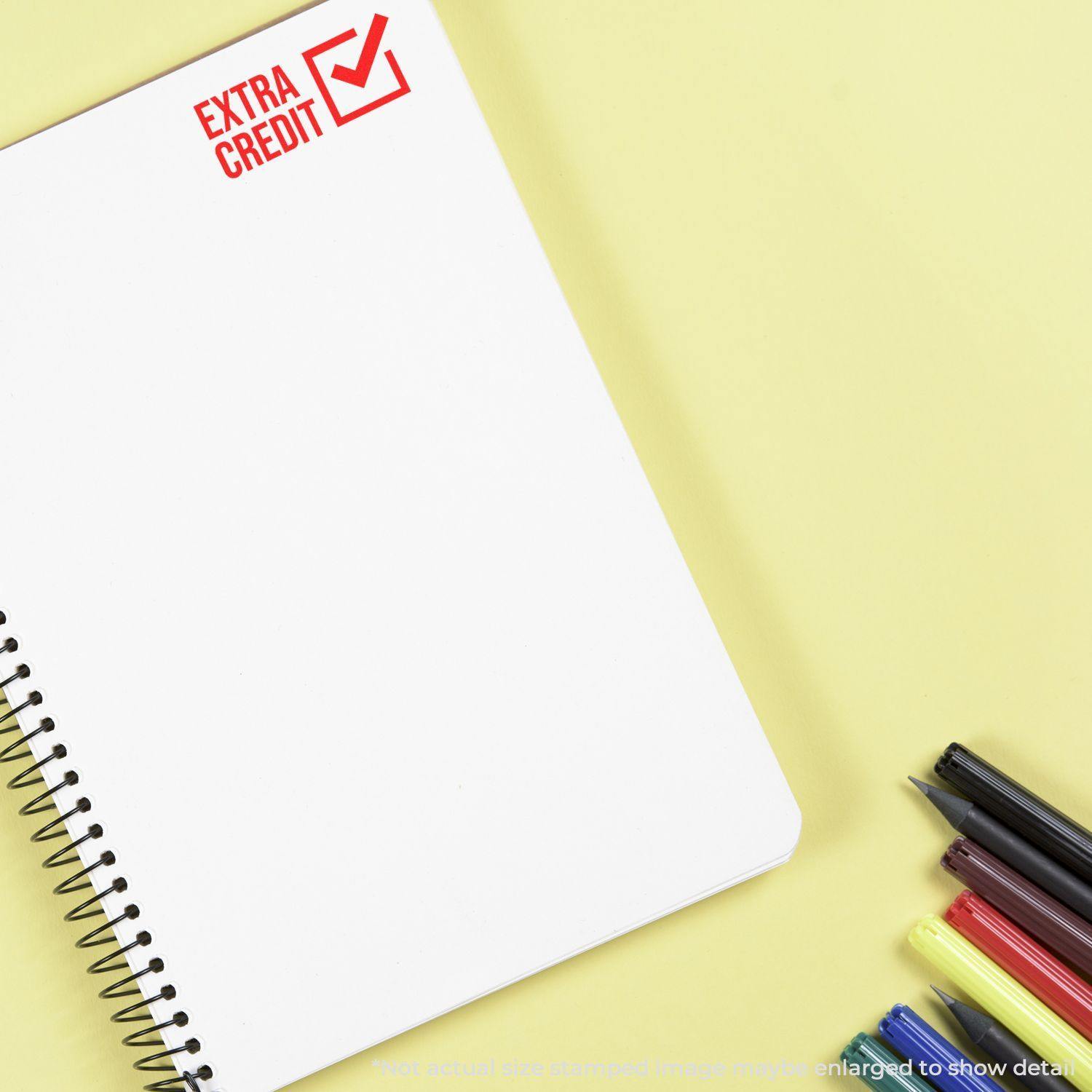 Slim Pre-Inked Extra Credit Stamp marking a notebook with Extra Credit in red ink, next to colorful pens on a yellow background.