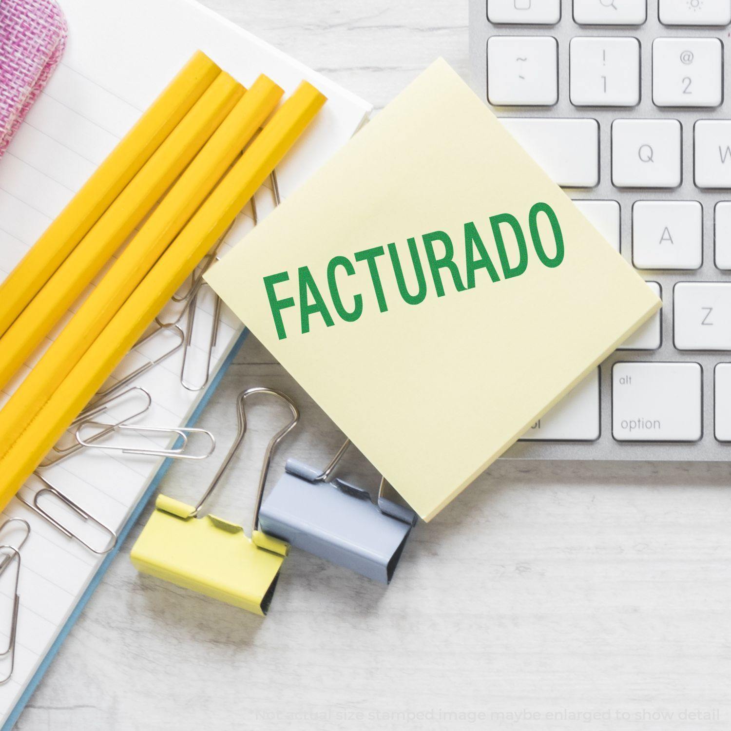 Facturado Rubber Stamp on a yellow sticky note, surrounded by pencils, paper clips, binder clips, and a keyboard on a desk.