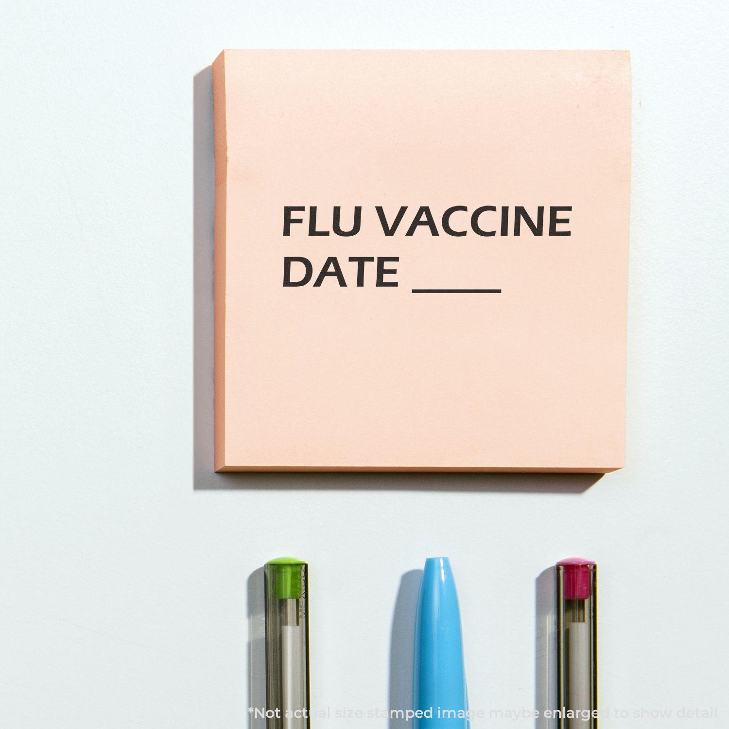 Flu Vaccine Date Rubber Stamp on a pink sticky note with blank space for date, placed above three pens with colored caps on a white surface.