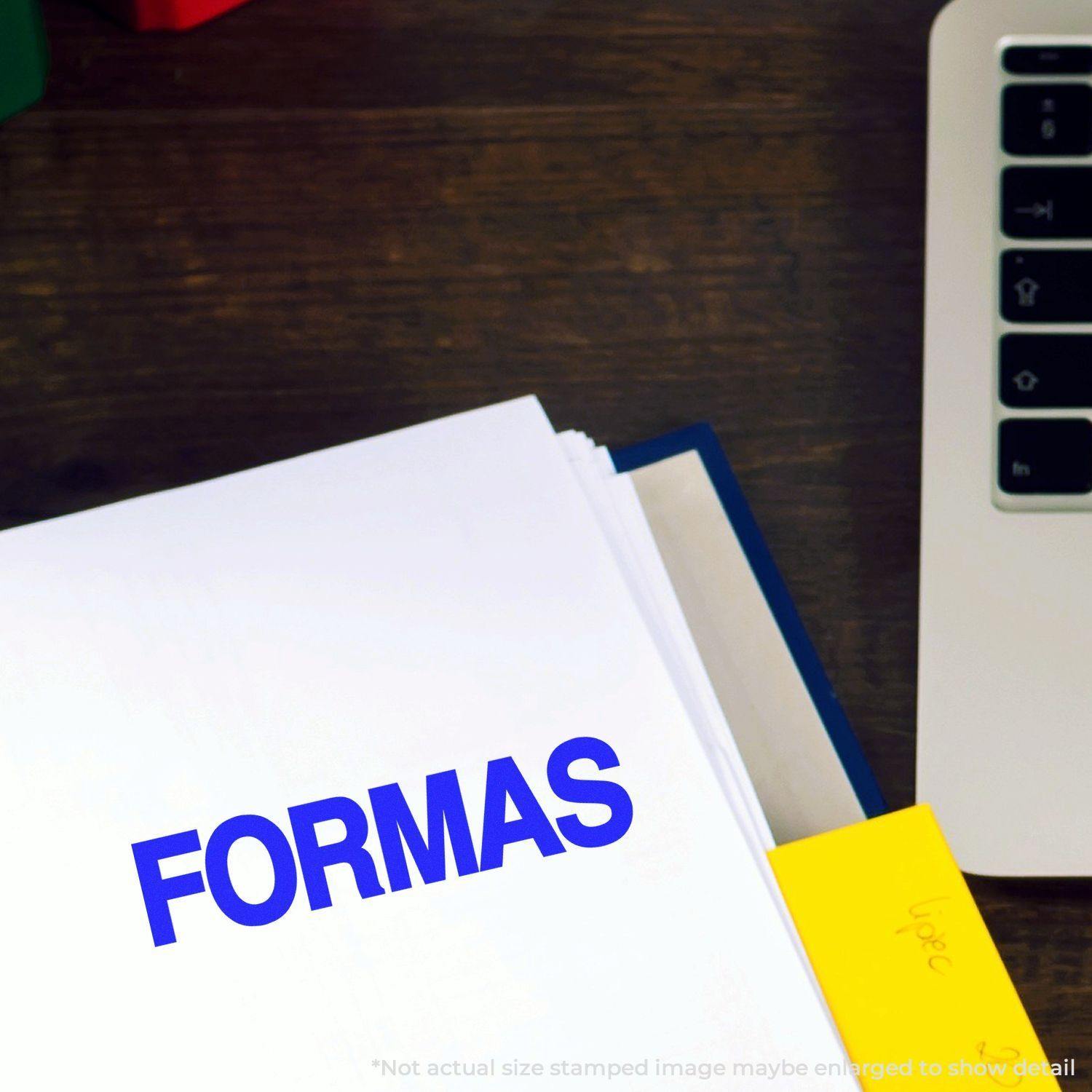 Large Pre-Inked Formas Stamp in use, stamping the word FORMAS in blue ink on a white document, next to a laptop on a wooden desk.