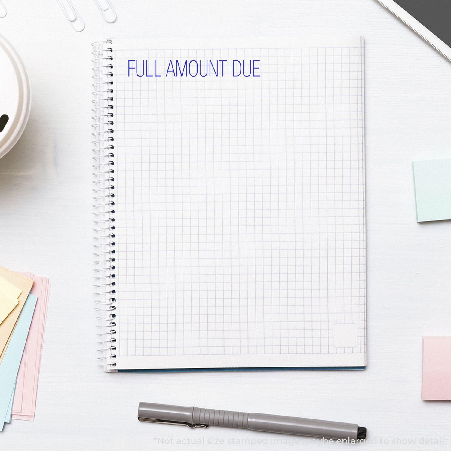 Large Full Amount Due Rubber Stamp imprint on a spiral notebook with grid paper, surrounded by office supplies on a white desk.