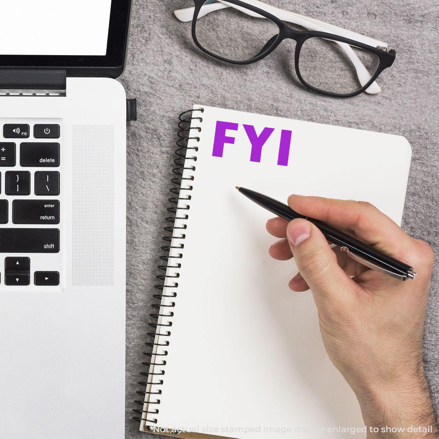 Hand holding a pen near a notebook with FYI stamped in purple using the FYI Rubber Stamp, next to a laptop and glasses.