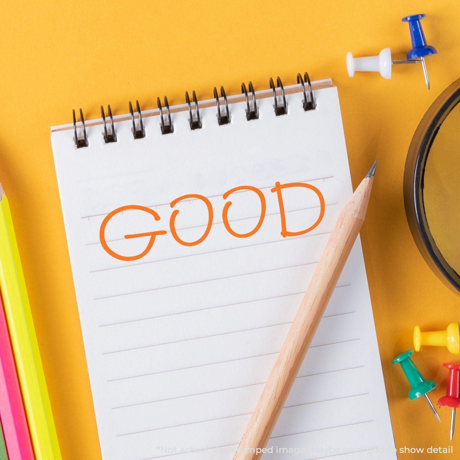 A notepad with GOOD stamped in orange using the Good Rubber Stamp, surrounded by colorful pencils, push pins, and a magnifying glass.