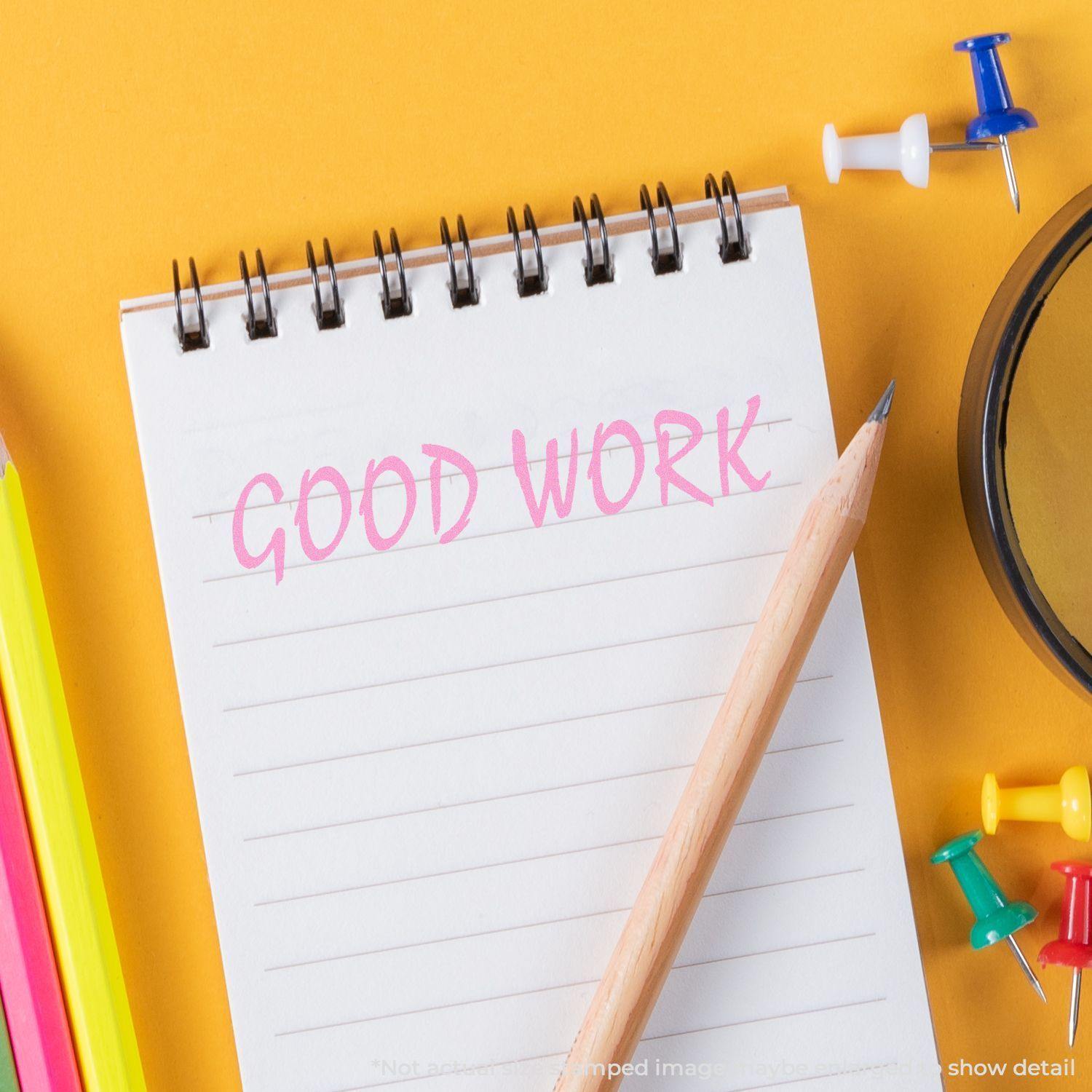 A Self Inking Good Work Stamp is used on a notebook with GOOD WORK stamped in pink. Nearby are colorful pencils, push pins, and a pencil.