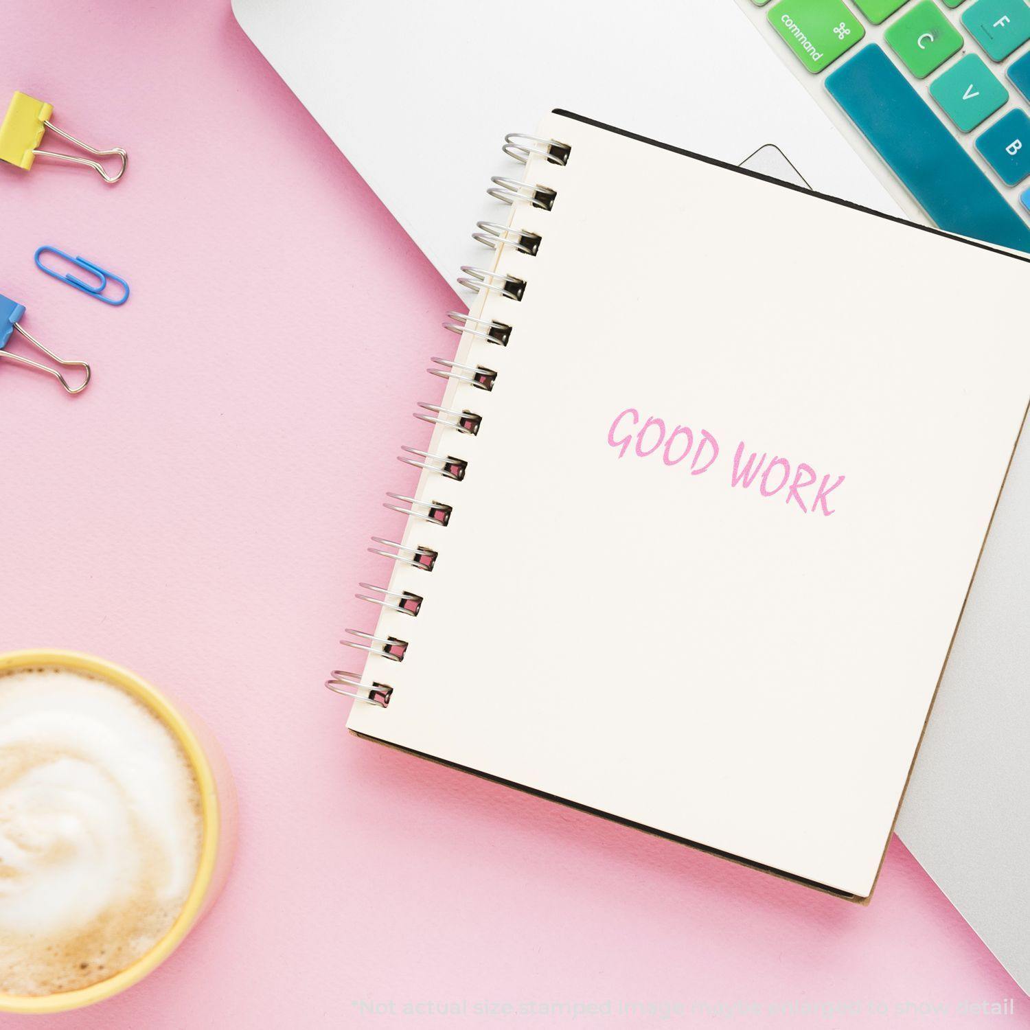 Large Pre-Inked Good Work Stamp used on a notebook, placed on a pink desk with a laptop, coffee, and colorful paper clips.