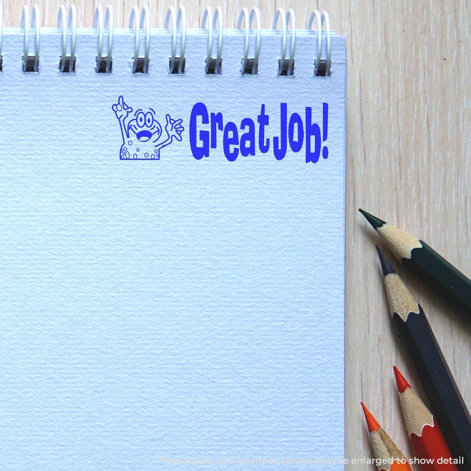 A notebook with a blue Great Job Rubber Stamp impression and three colored pencils (black, green, and red) on a wooden surface.