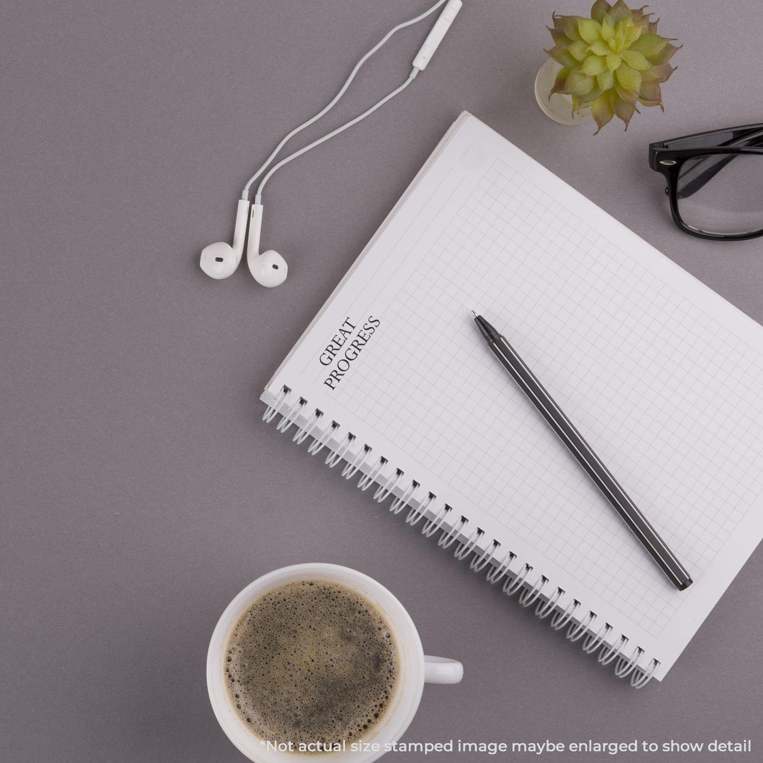 A notebook stamped with GREAT PROGRESS using the Large Pre-Inked Great Progress Stamp, surrounded by a pen, coffee, glasses, and earphones.