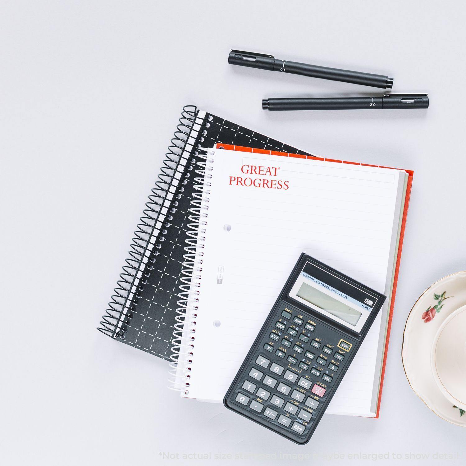 Self Inking Great Progress Stamp on a notebook with a calculator, two pens, and a teacup on a white surface.