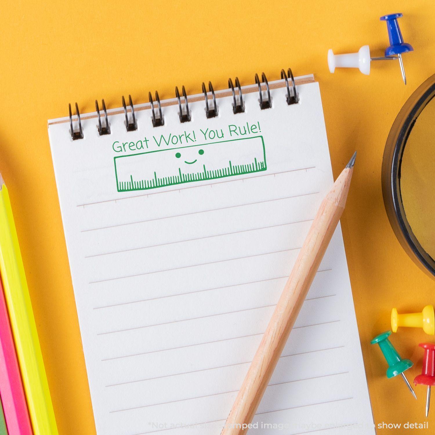 Large Self Inking Great Work You Rule Stamp on a notepad with a pencil, colorful pens, push pins, and a magnifying glass nearby.