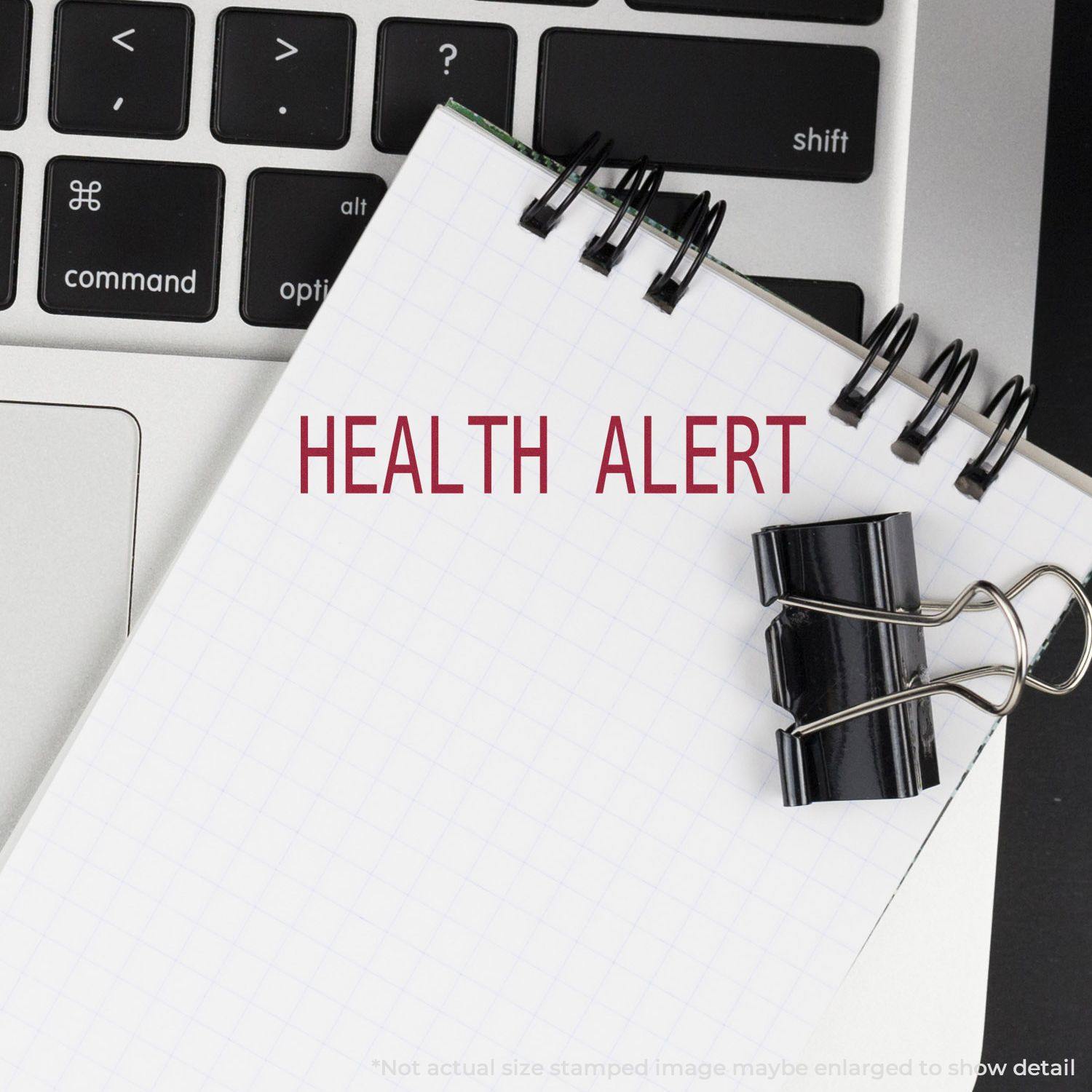 A Large Health Alert Rubber Stamp imprint on a spiral notebook page, placed on a laptop keyboard with a black binder clip.