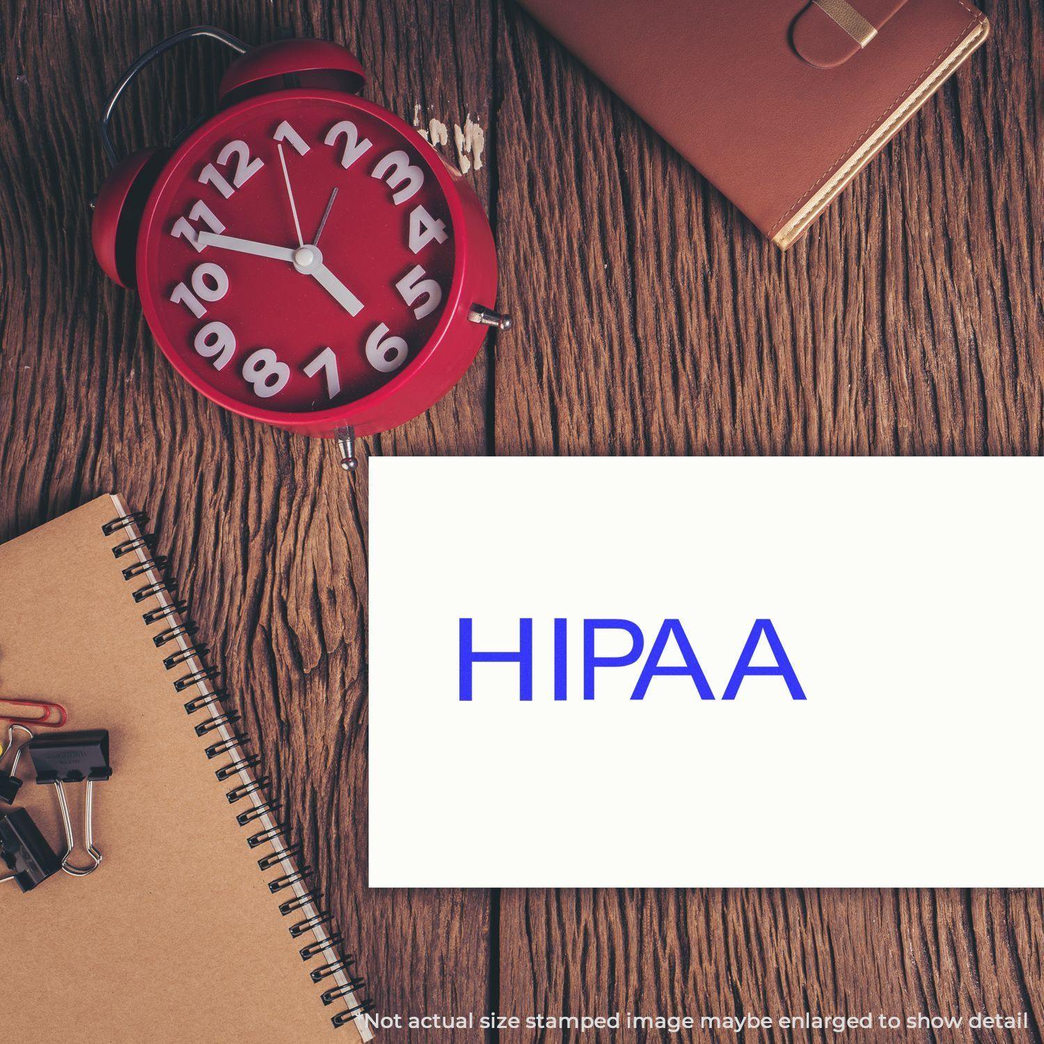 Large Pre-Inked HIPAA Stamp on a white card, placed on a wooden desk with a red clock, notebook, and office supplies.