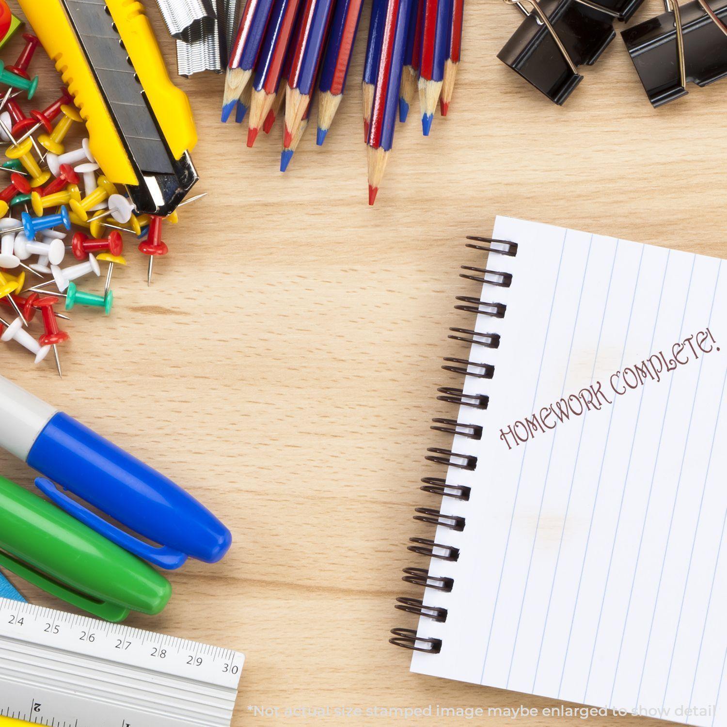 Large Homework Complete Rubber Stamp used on a spiral notebook, surrounded by colorful stationery items like pens, push pins, and clips.