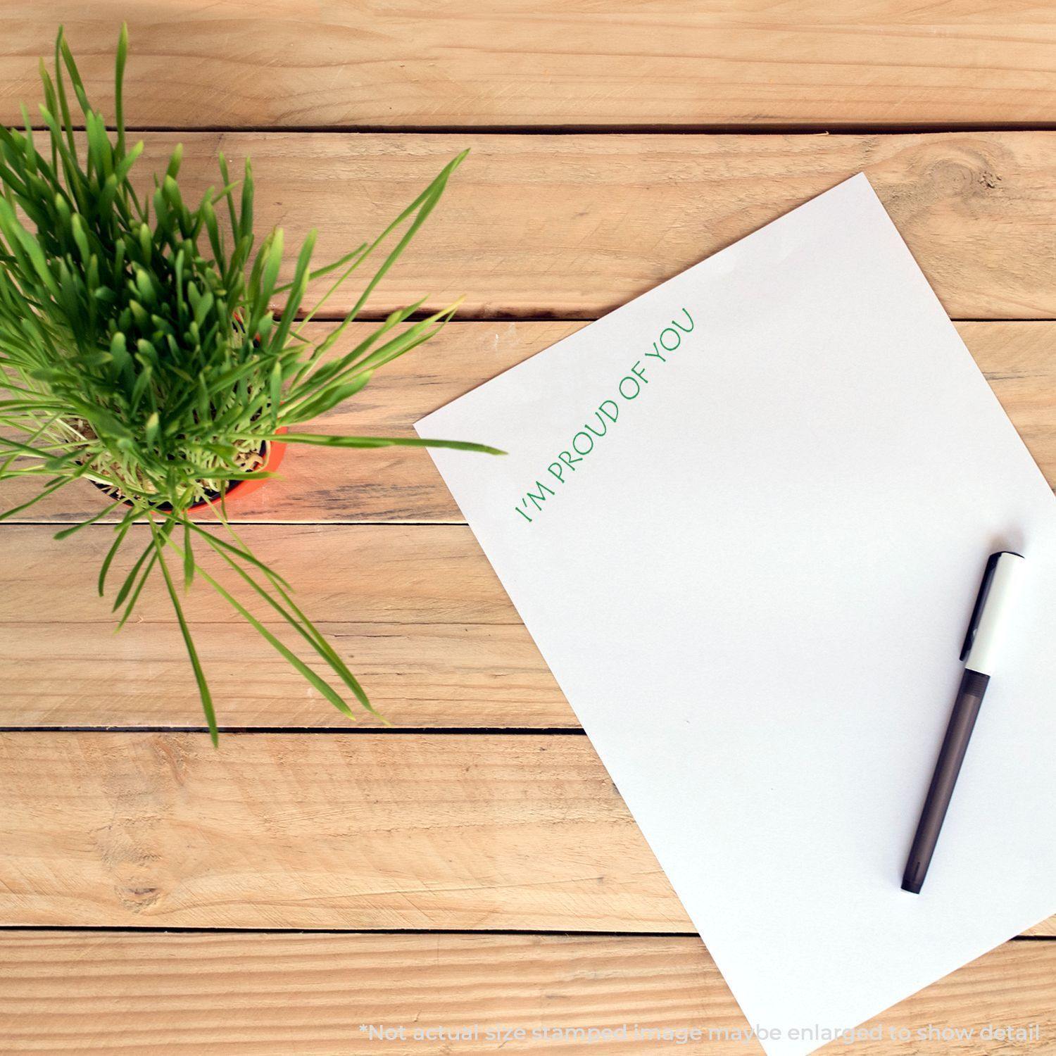 A Large Self Inking I'm Proud Of You Stamp used on a white sheet of paper, placed on a wooden table next to a green plant and a pen.