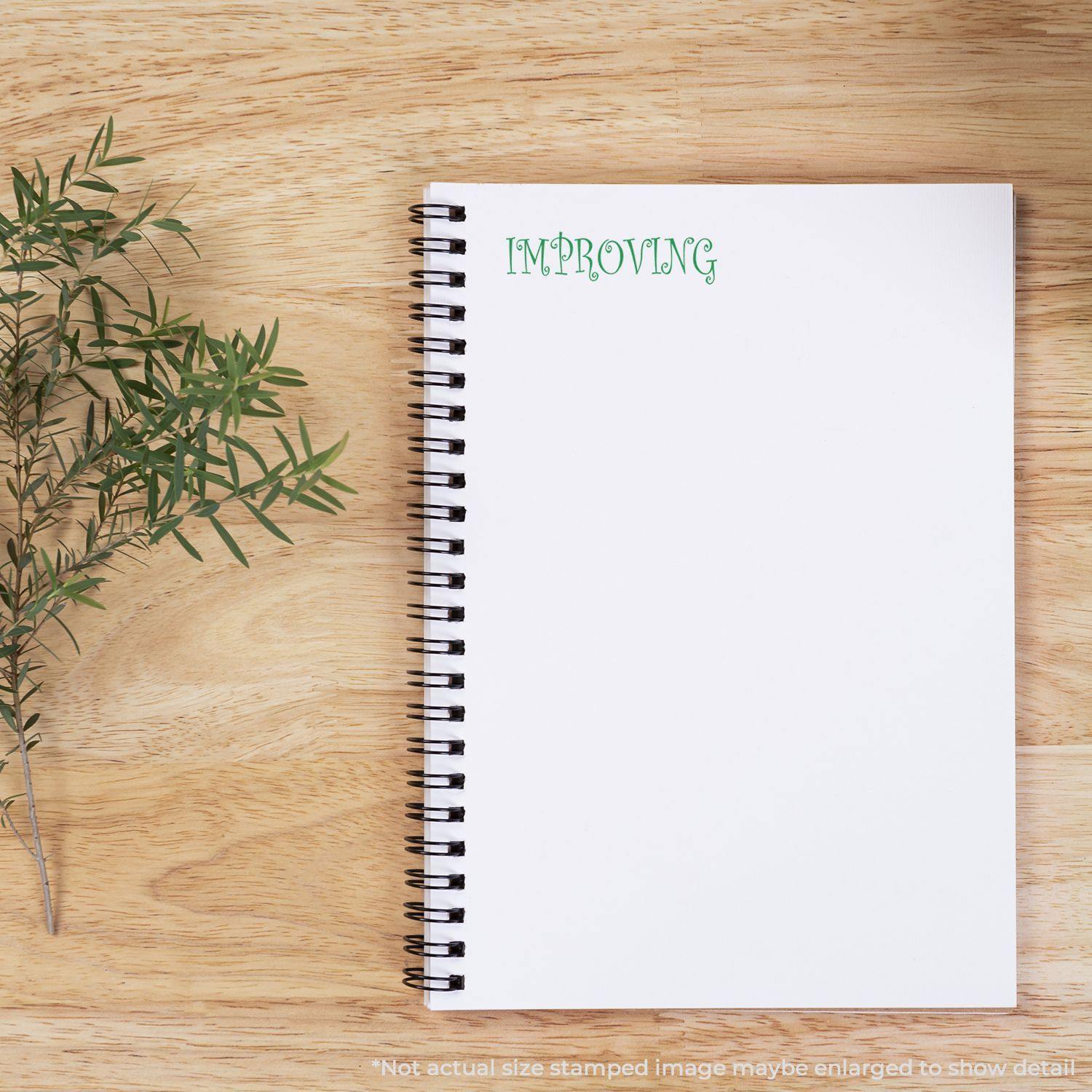 A notebook with IMPROVING stamped in green using the Large Pre-Inked Improving Stamp, placed on a wooden surface with a sprig of greenery.