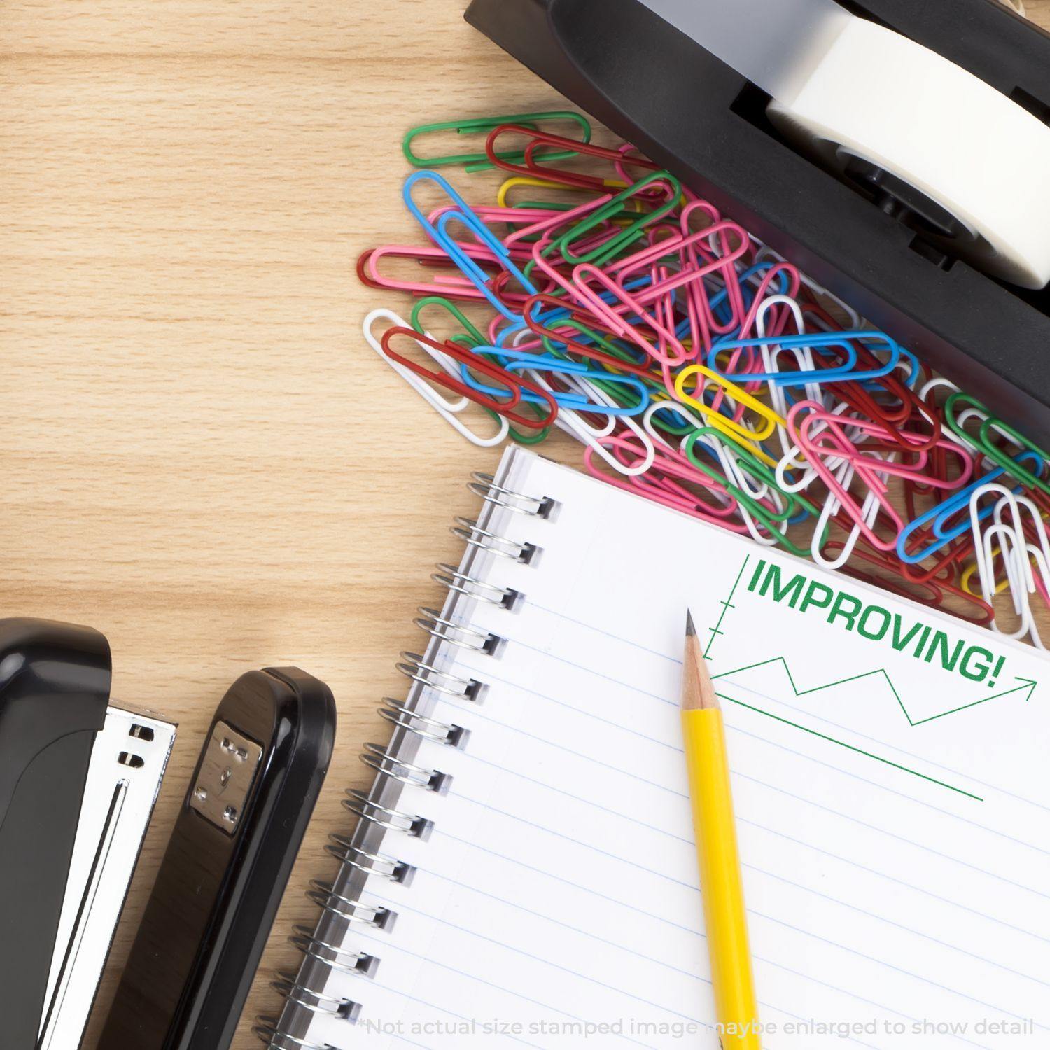 Improving with Chart Icon Rubber Stamp on a notebook, surrounded by a pencil, stapler, tape dispenser, and colorful paperclips on a wooden desk.