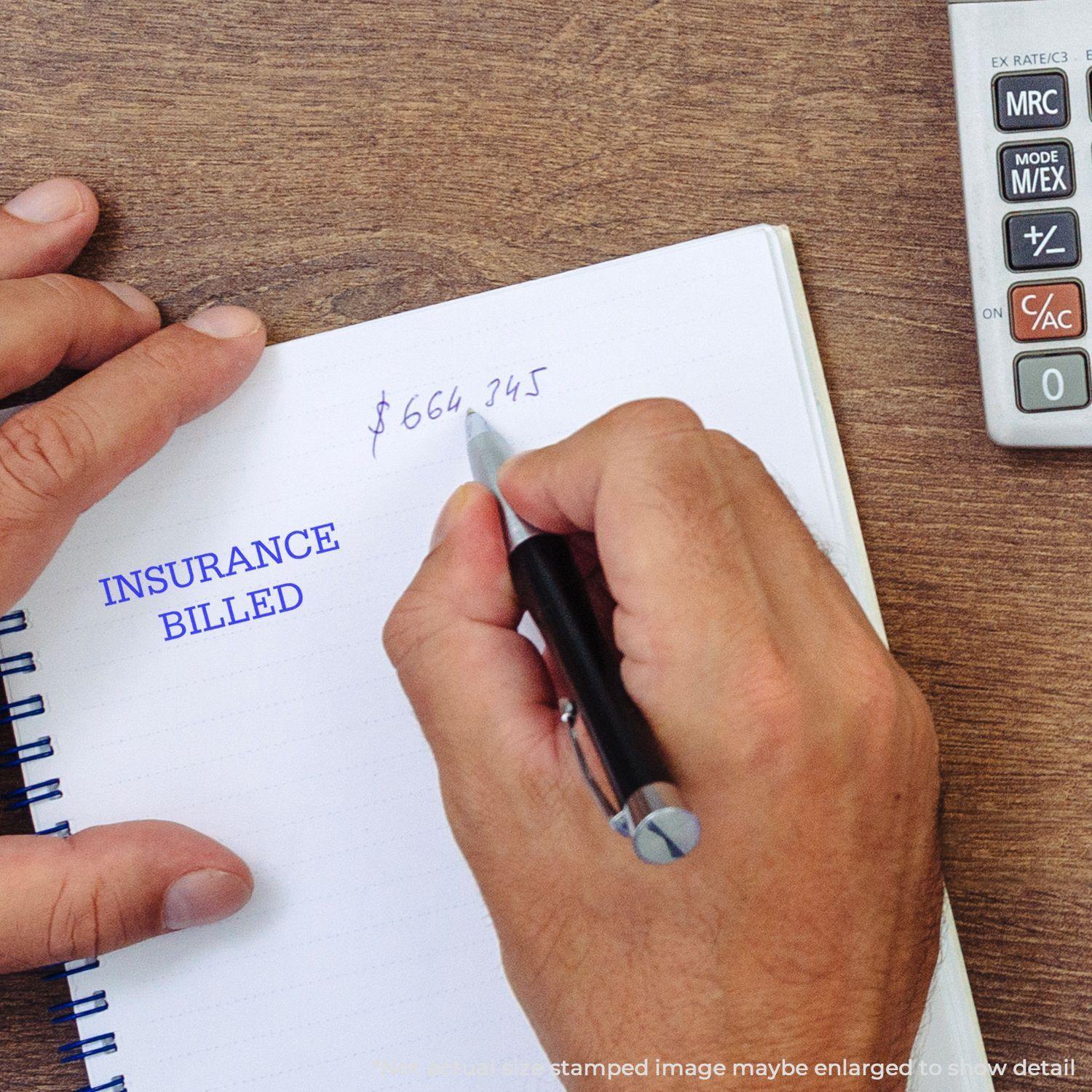 A hand writing in a notebook with Insurance Billed stamped in blue ink, next to a calculator. Product name: Insurance Billed Rubber Stamp.