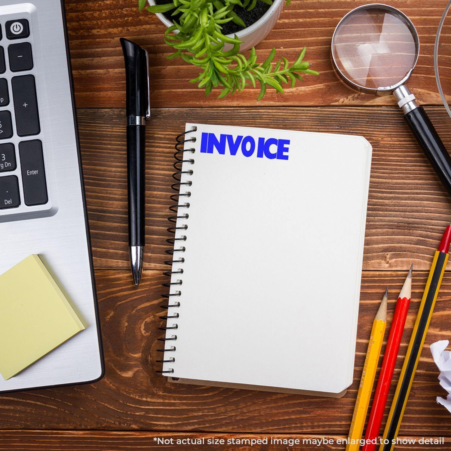 Slim Pre-Inked Invoice Stamp on a spiral notebook, surrounded by office supplies including a laptop, pen, magnifying glass, and plant.