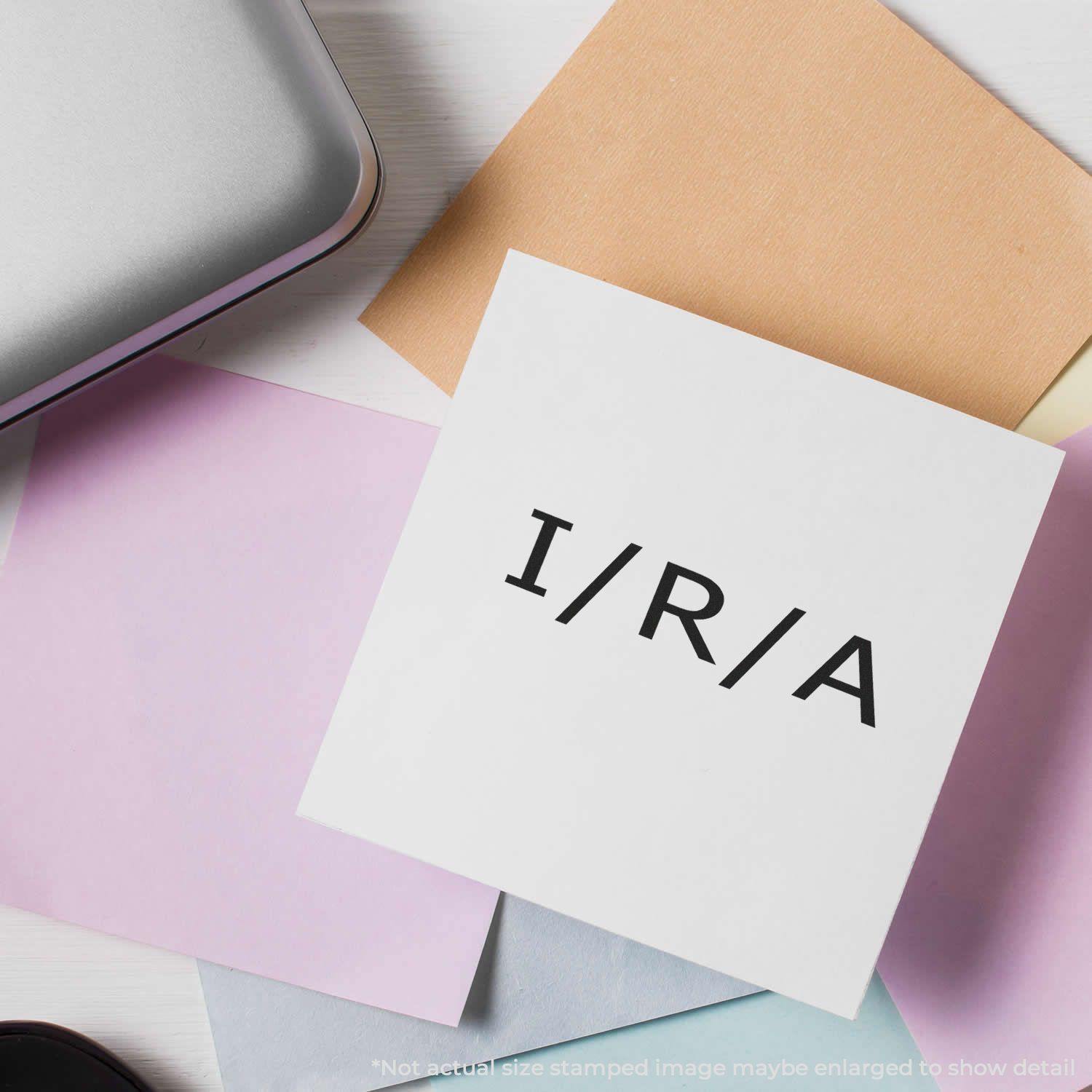 A white card stamped with I/R/A using the I/R/A Rubber Stamp, surrounded by pastel-colored papers and a silver case.