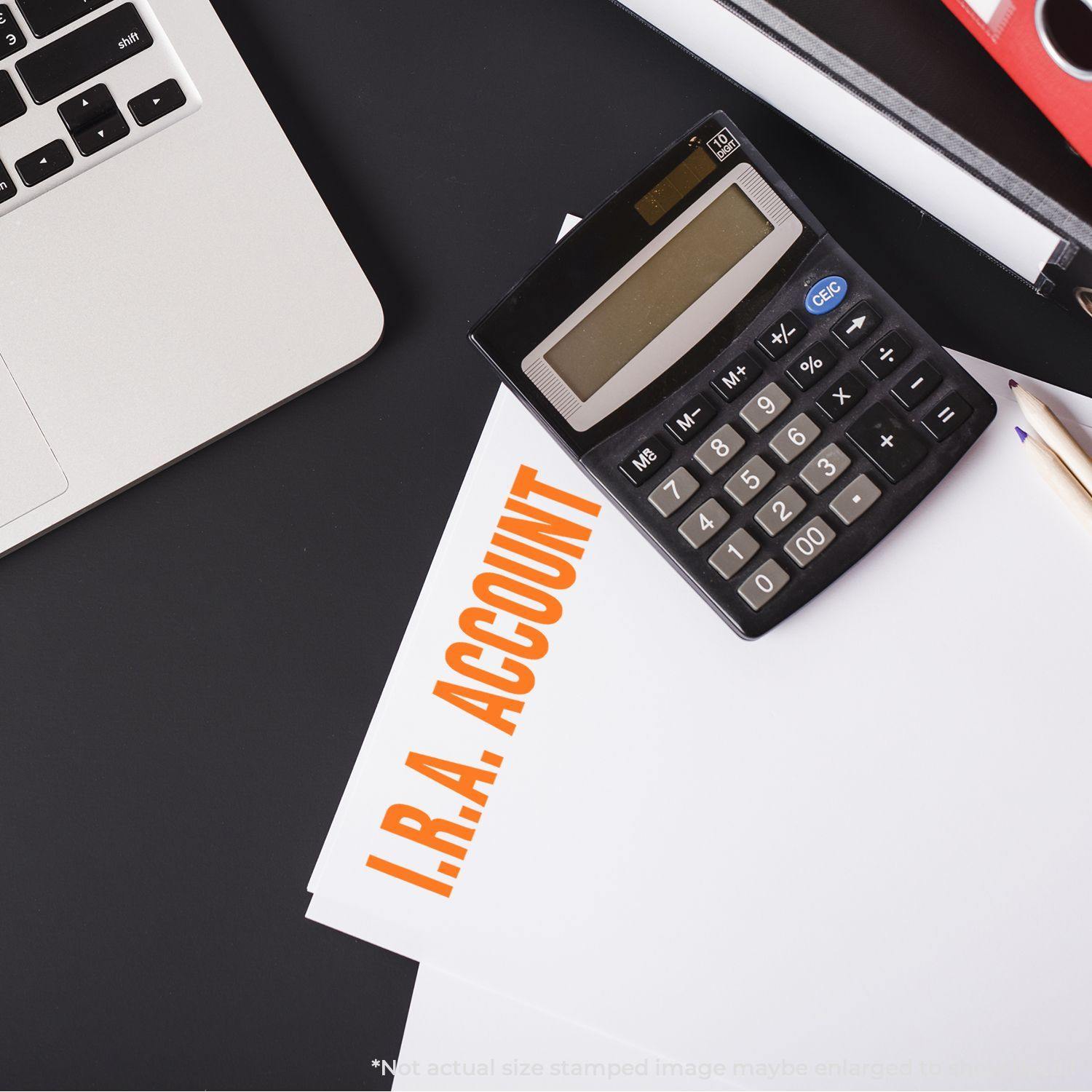 A Large Pre-Inked I.R.A. Account Stamp is used on a document next to a calculator, laptop, and pencil on a black desk.