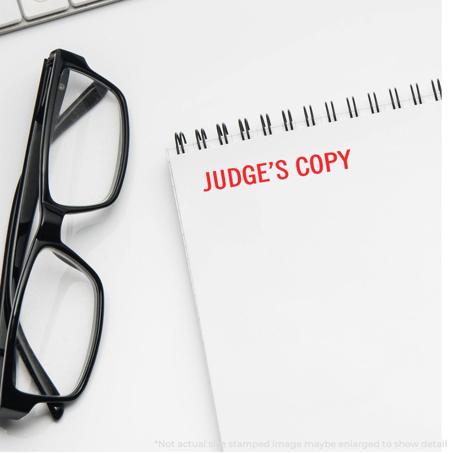 A notebook with Judge's Copy stamped in red, next to black glasses and a keyboard. Judge's Copy Rubber Stamp used on the notebook.