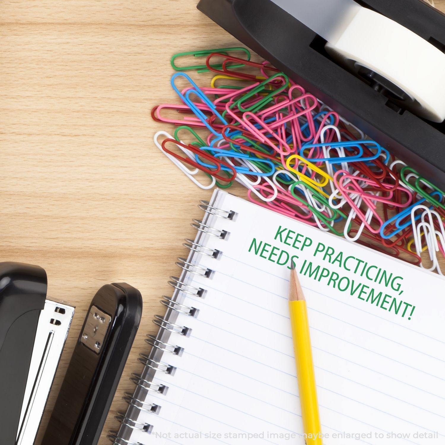 A desk with a stapler, tape dispenser, colorful paperclips, and a notebook stamped with Large Keep Practicing Needs Improvement Rubber Stamp in green.