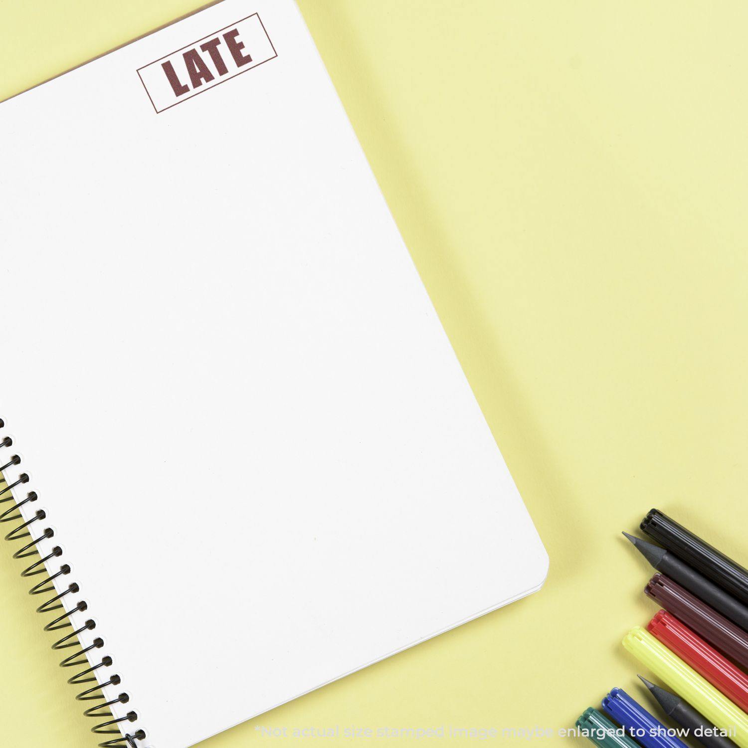 A Large Pre-Inked Late with Border Stamp marks LATE on a white spiral notebook, with pens and highlighters on a yellow background.