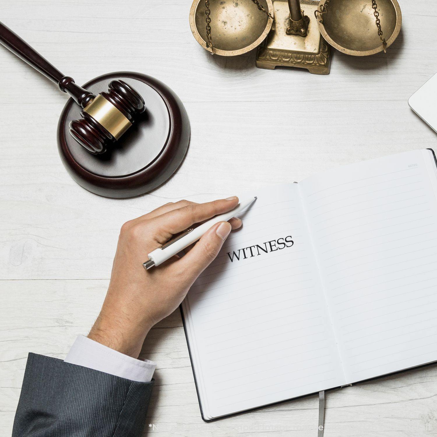 A hand holding a pen near a notebook with WITNESS written, a gavel, and scales of justice, featuring a Slim Pre-Inked Witness Stamp.