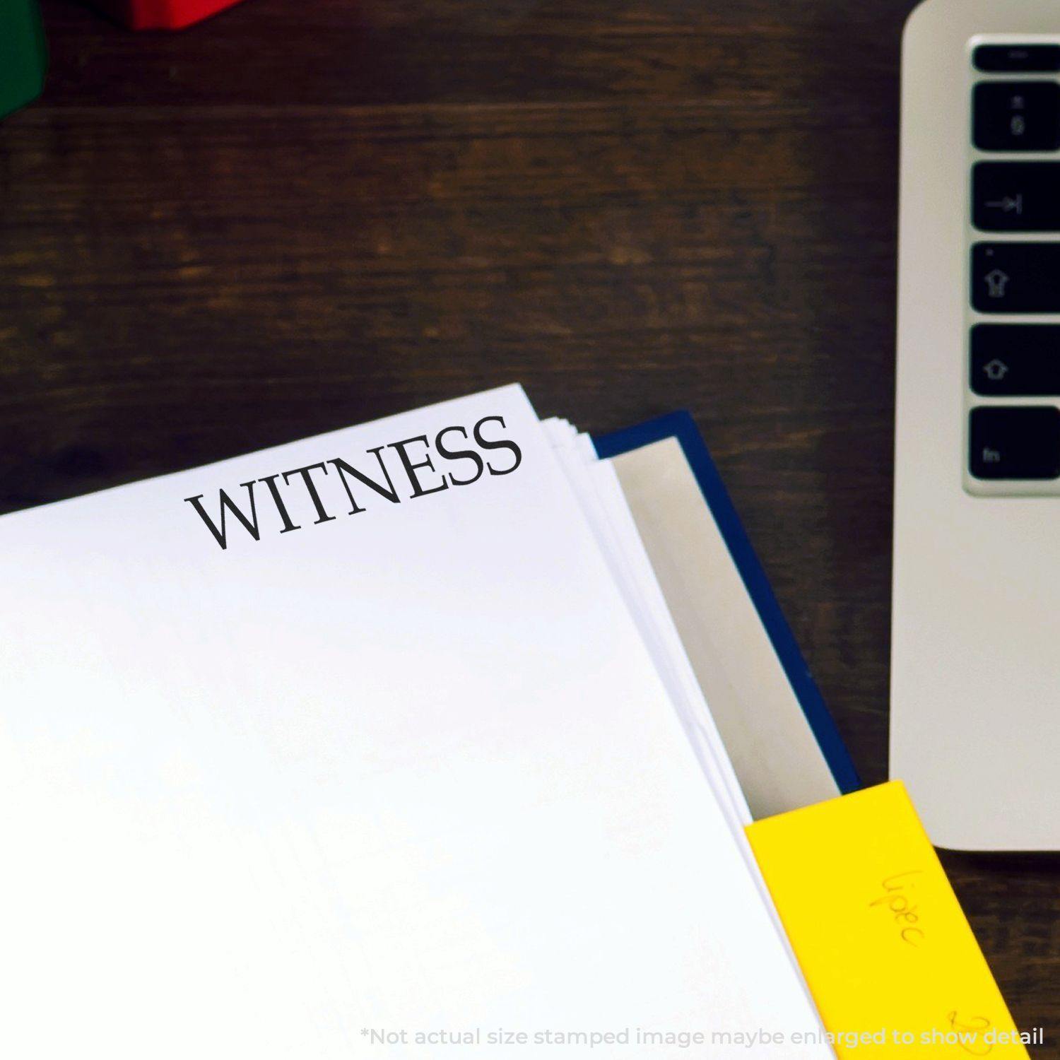 Large Witness Rubber Stamp imprint on a white document next to a laptop and a yellow sticky note on a wooden desk.