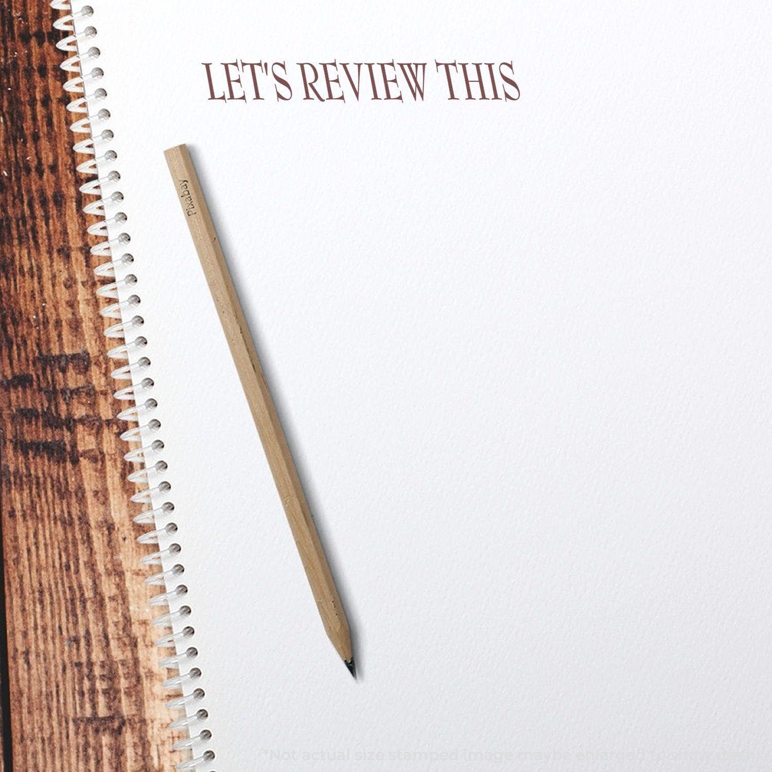 Large Lets Review This Rubber Stamp impression on white paper with a pencil beside it on a wooden surface.