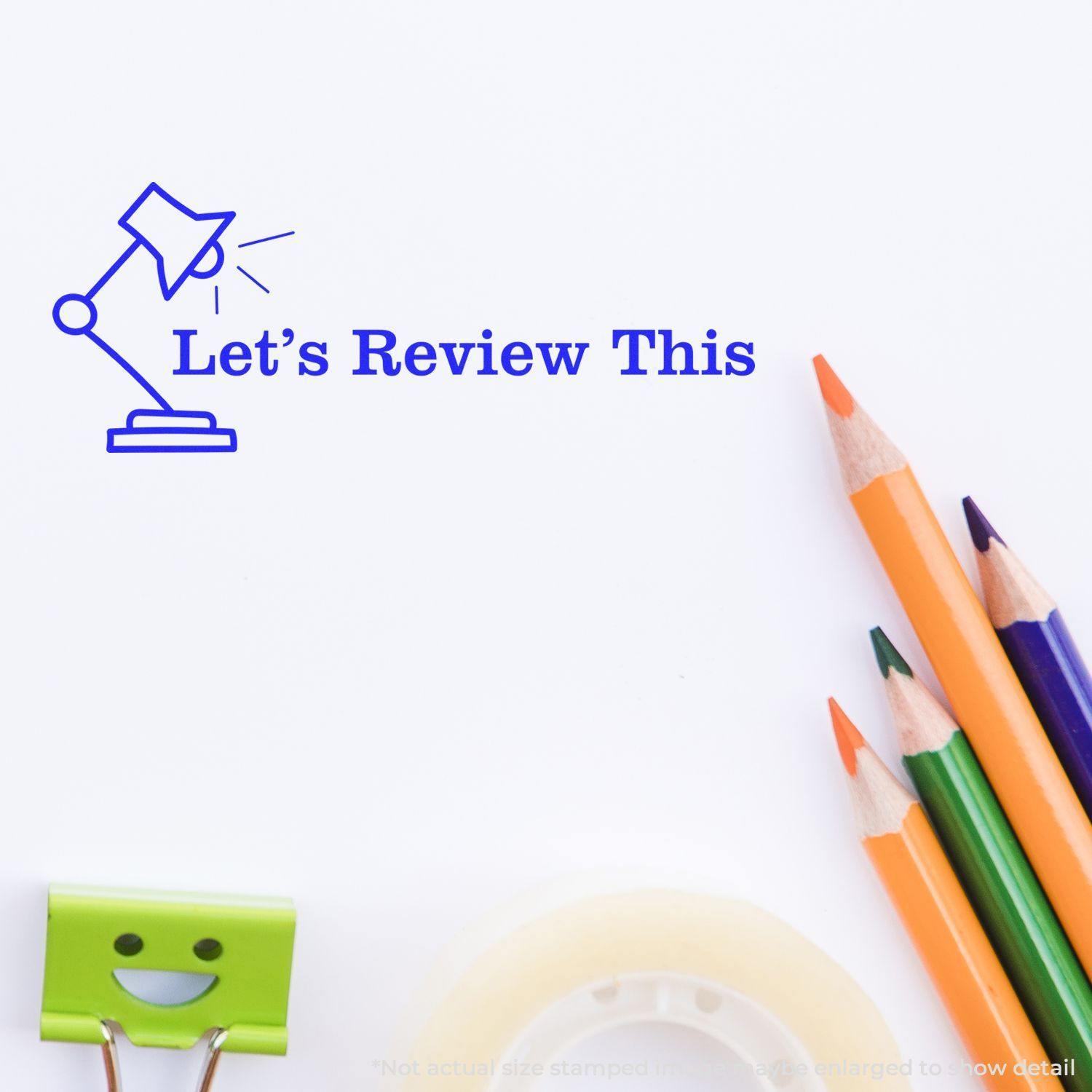 Large Lets Review This with Lamp Rubber Stamp in blue ink on white paper, surrounded by colorful pencils, a smiling clip, and tape.