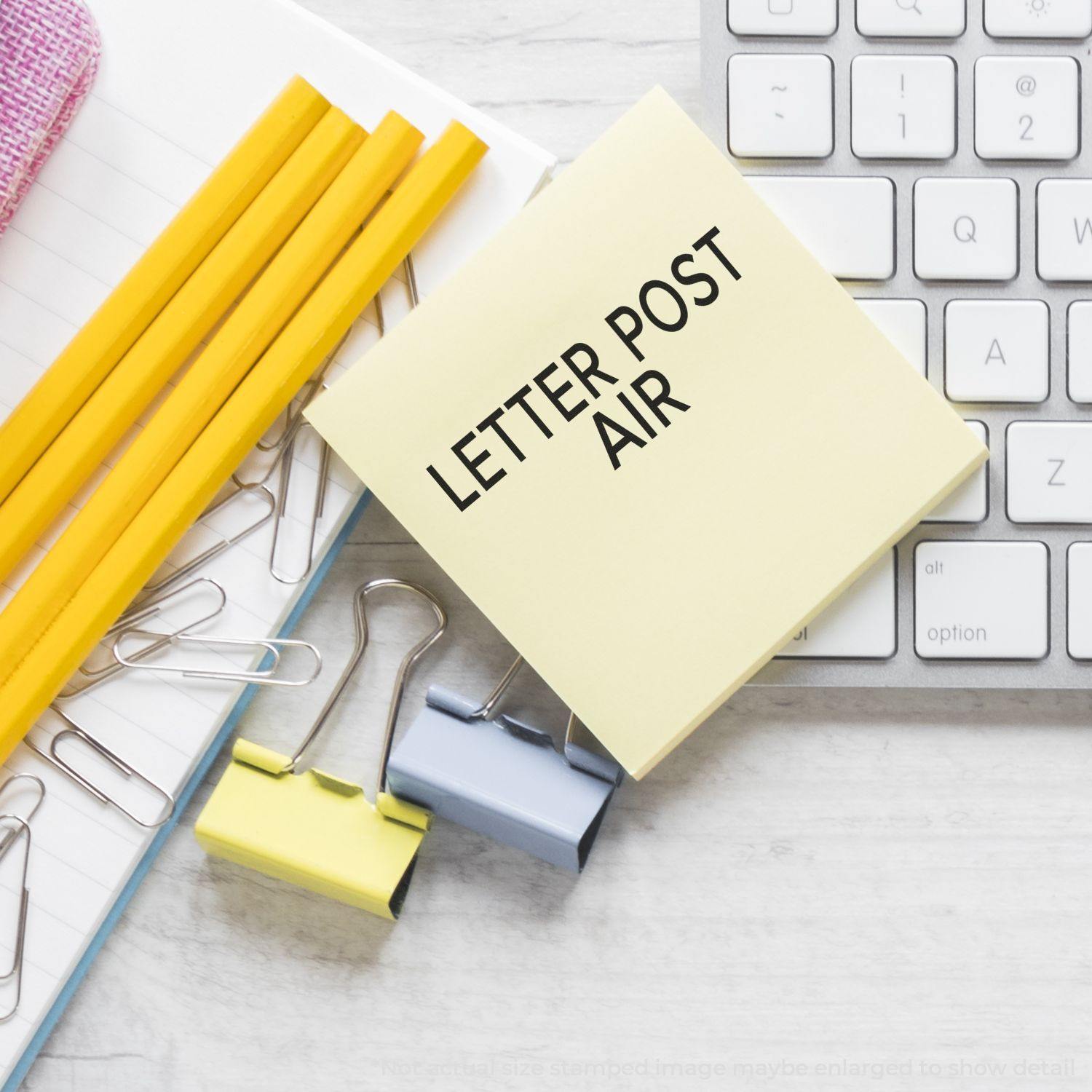 Large Pre-Inked Letter Post Air Stamp on a yellow sticky note, surrounded by pencils, paper clips, and a keyboard.