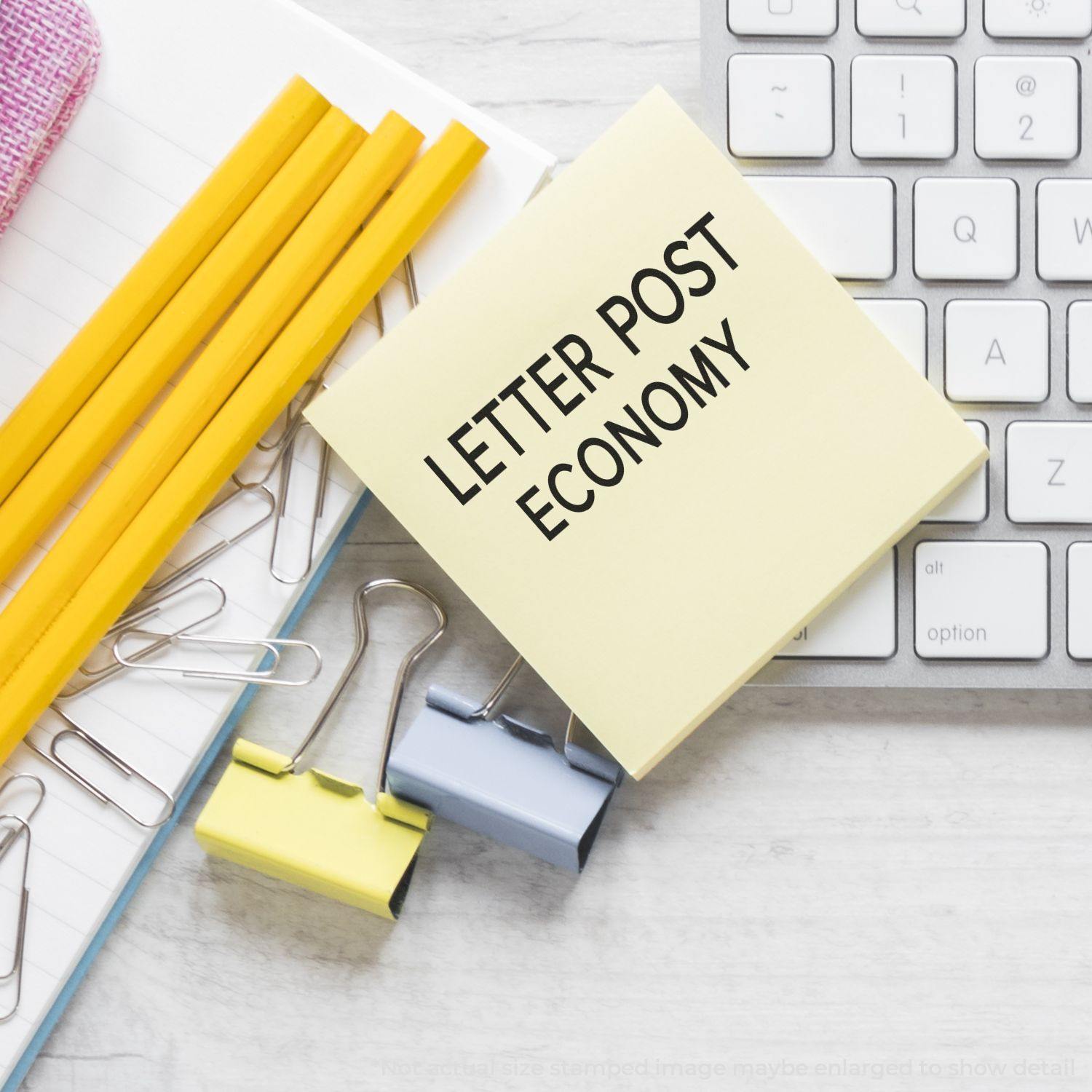 Slim Pre-Inked Letter Post Economy Stamp on a sticky note beside pencils, paper clips, and a keyboard on a desk.