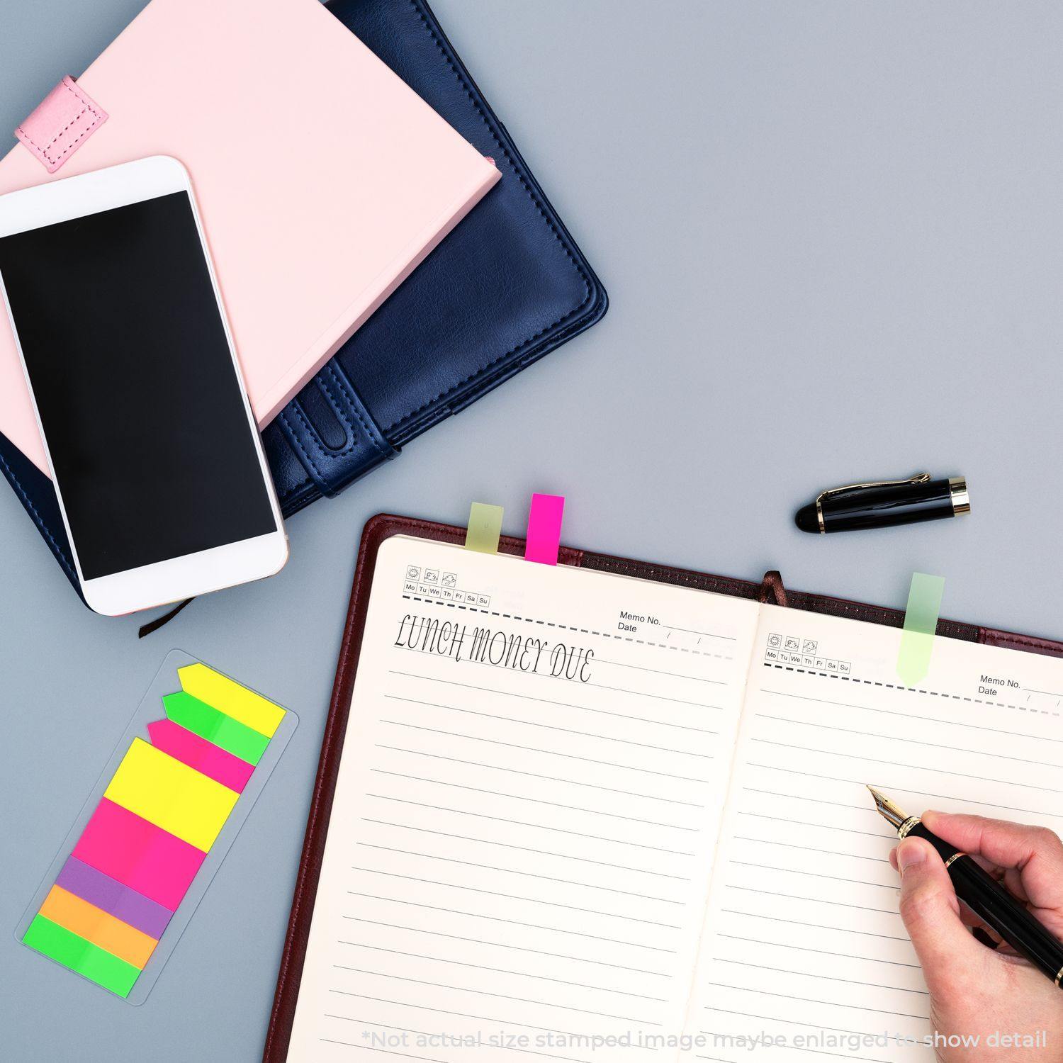 Person using a Slim Pre-Inked Lunch Money Due School Stamp on a notebook, with a phone, sticky notes, and pen nearby.