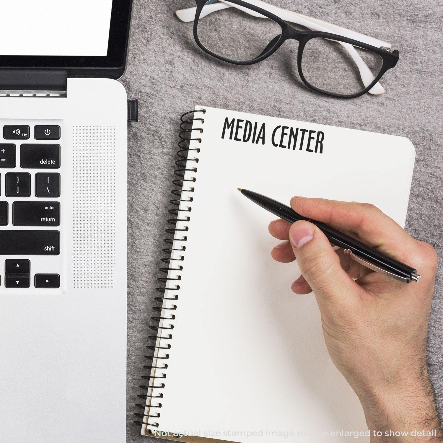 Hand using Large Self Inking Media Center Stamp on a notebook, with a laptop and glasses nearby.