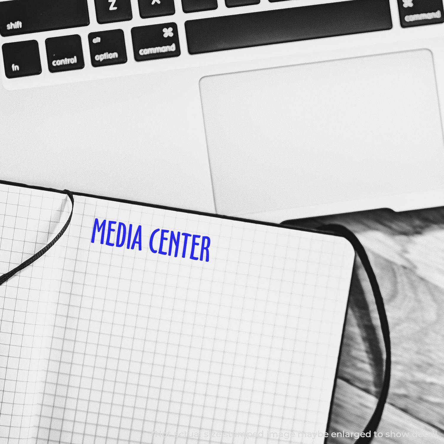 A Large Self Inking Media Center Stamp marks MEDIA CENTER in blue ink on a grid notebook next to a laptop keyboard.