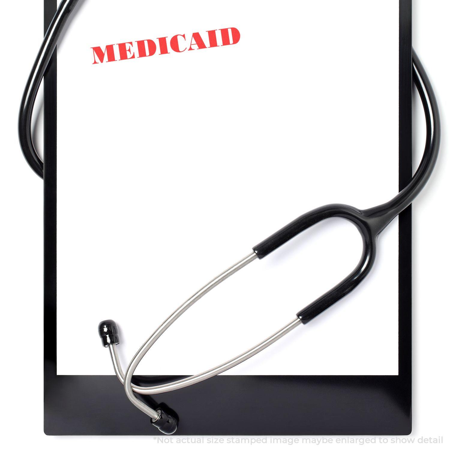 A Large Medicaid Rubber Stamp marks MEDICAID in red on a white paper clipped to a black clipboard with a stethoscope draped over it.