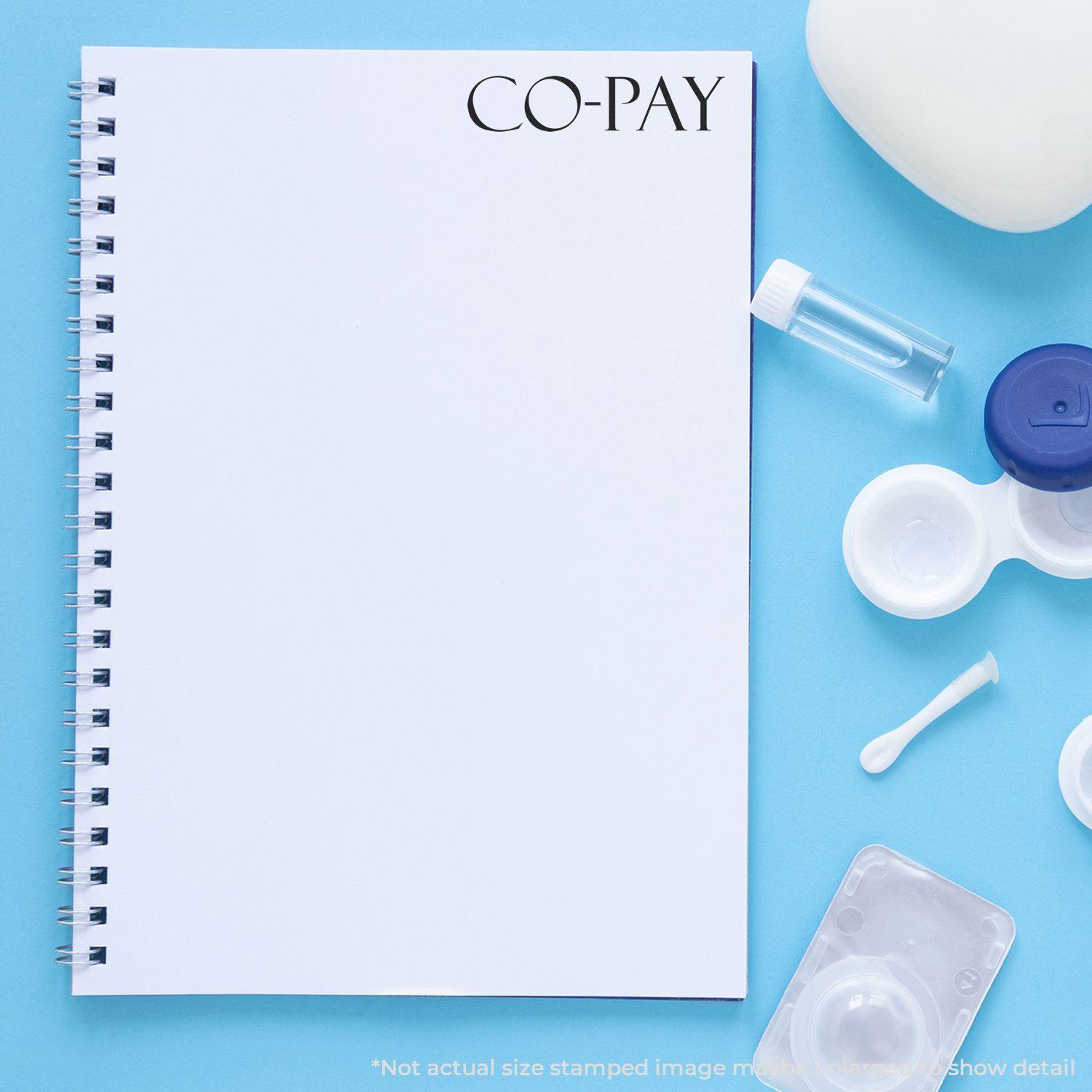 A Large Co Pay Rubber Stamp is used on a blank notebook page, surrounded by medical items like a contact lens case, solution bottle, and cotton swab.