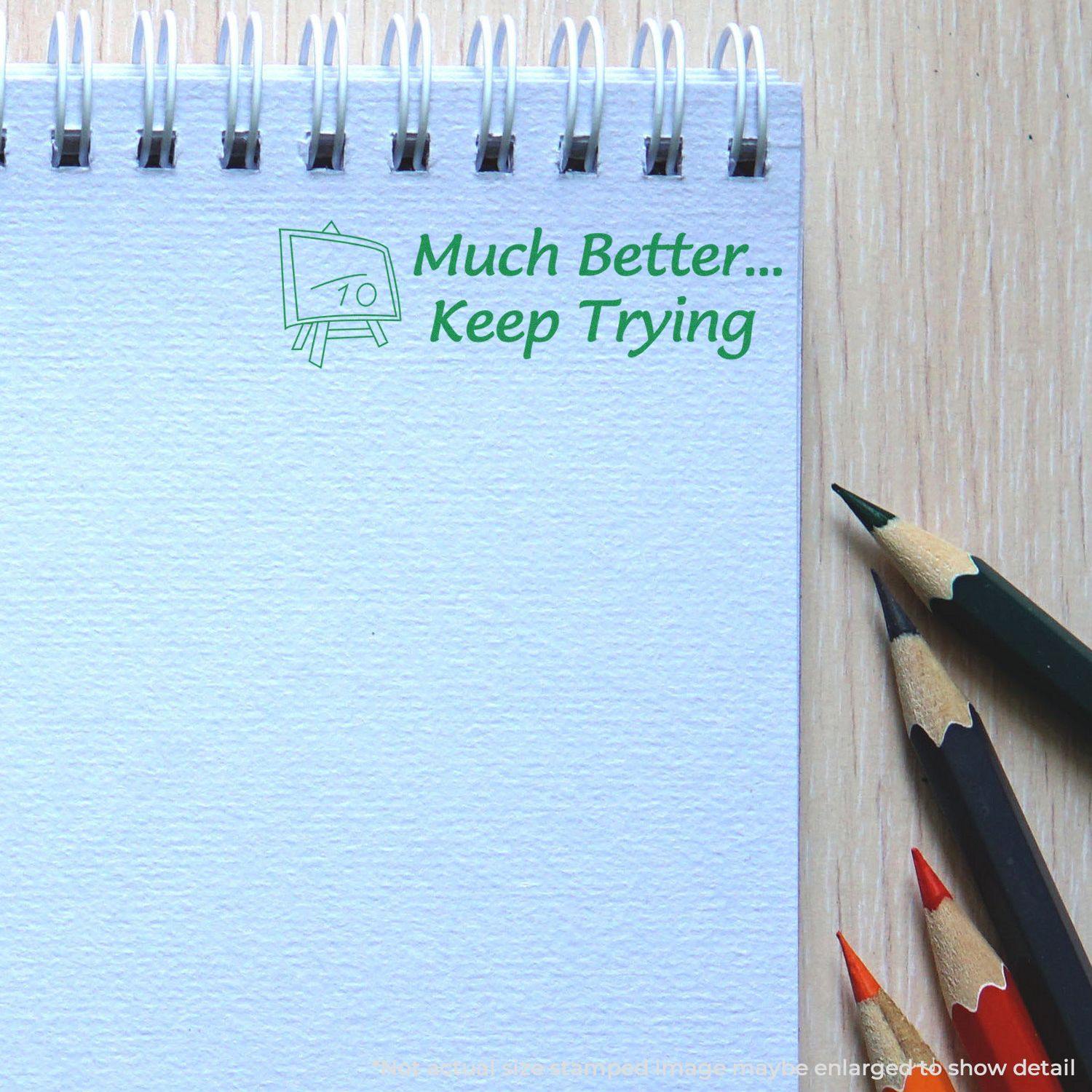 A notebook with a Large Much Better Keep Trying Rubber Stamp impression in green ink, next to colored pencils on a wooden surface.