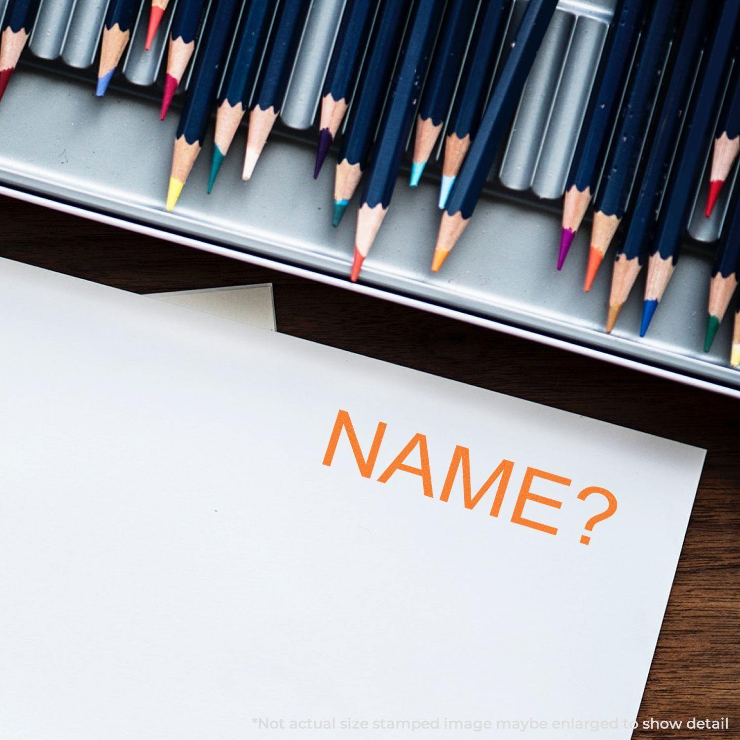 Large Pre-Inked Name Stamp in use, stamping NAME? in orange ink on white paper, with colored pencils in the background.