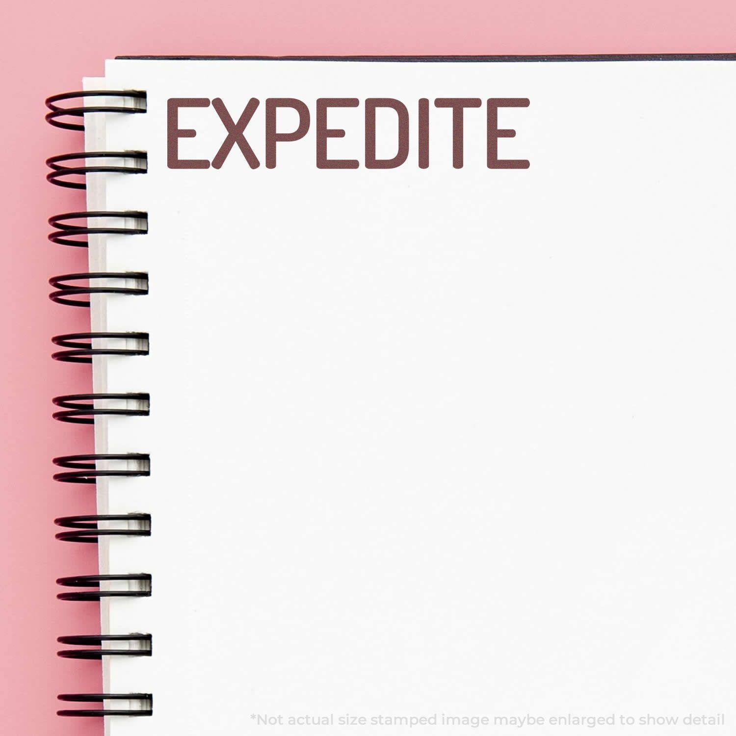 A Large Narrow Expedite Rubber Stamp imprint on a spiral-bound notebook with a pink background.