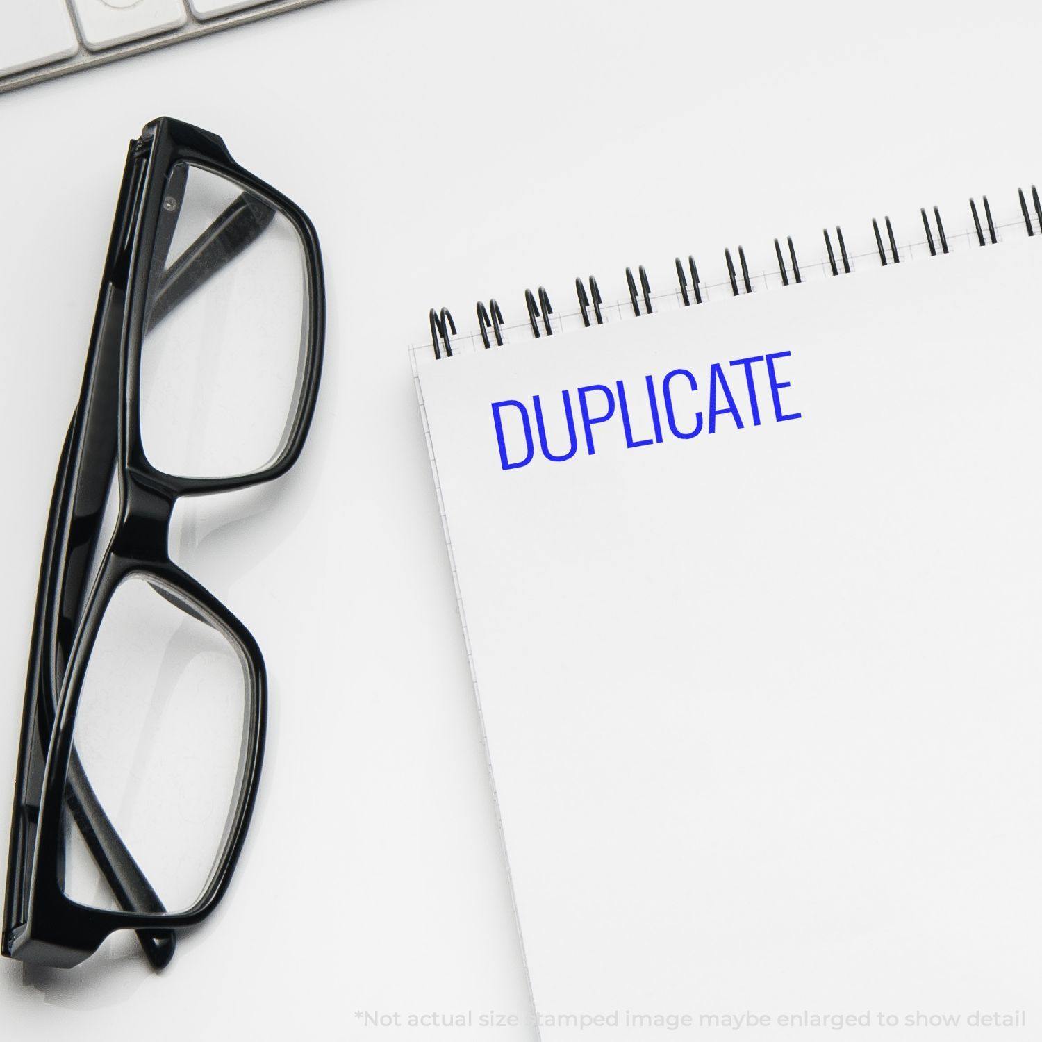 Large Self Inking Narrow Font Duplicate Stamp marking DUPLICATE in blue on a white notepad, next to a pair of black glasses.