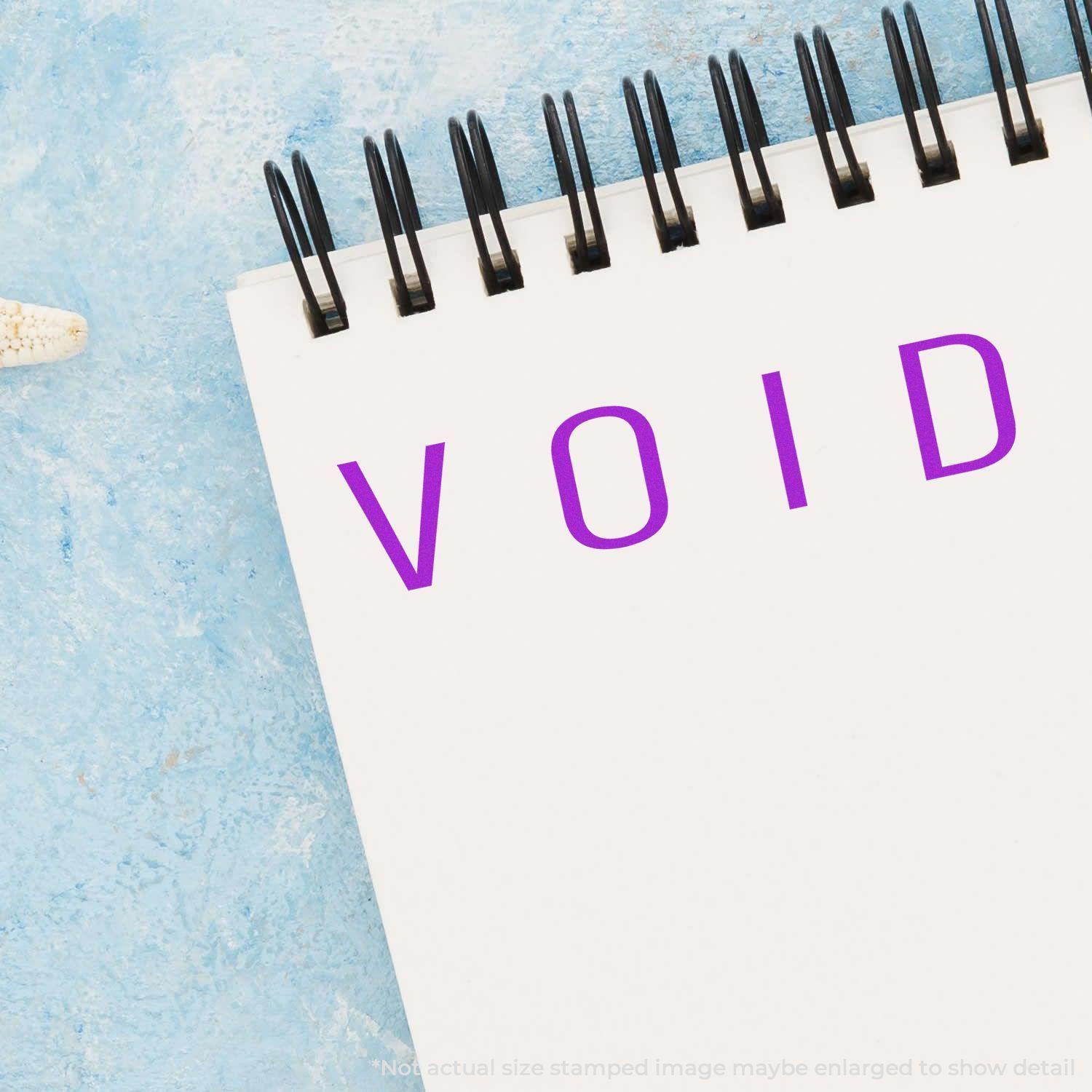 Slim Pre-Inked Narrow Void Stamp used on a white notepad with a blue background, showing the word VOID in purple ink.