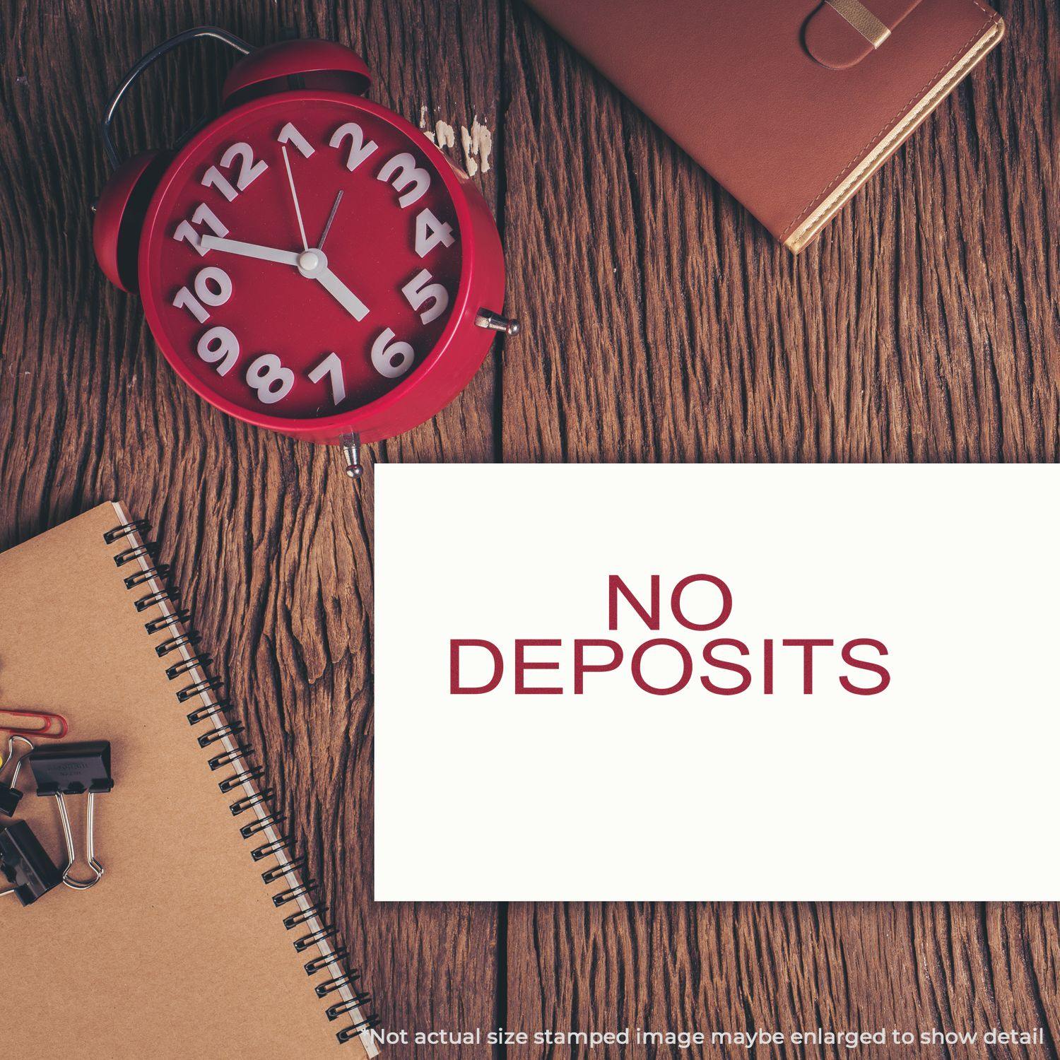 Large No Deposits Rubber Stamp used on white paper, placed on a wooden desk with a red clock, notebook, and office supplies.