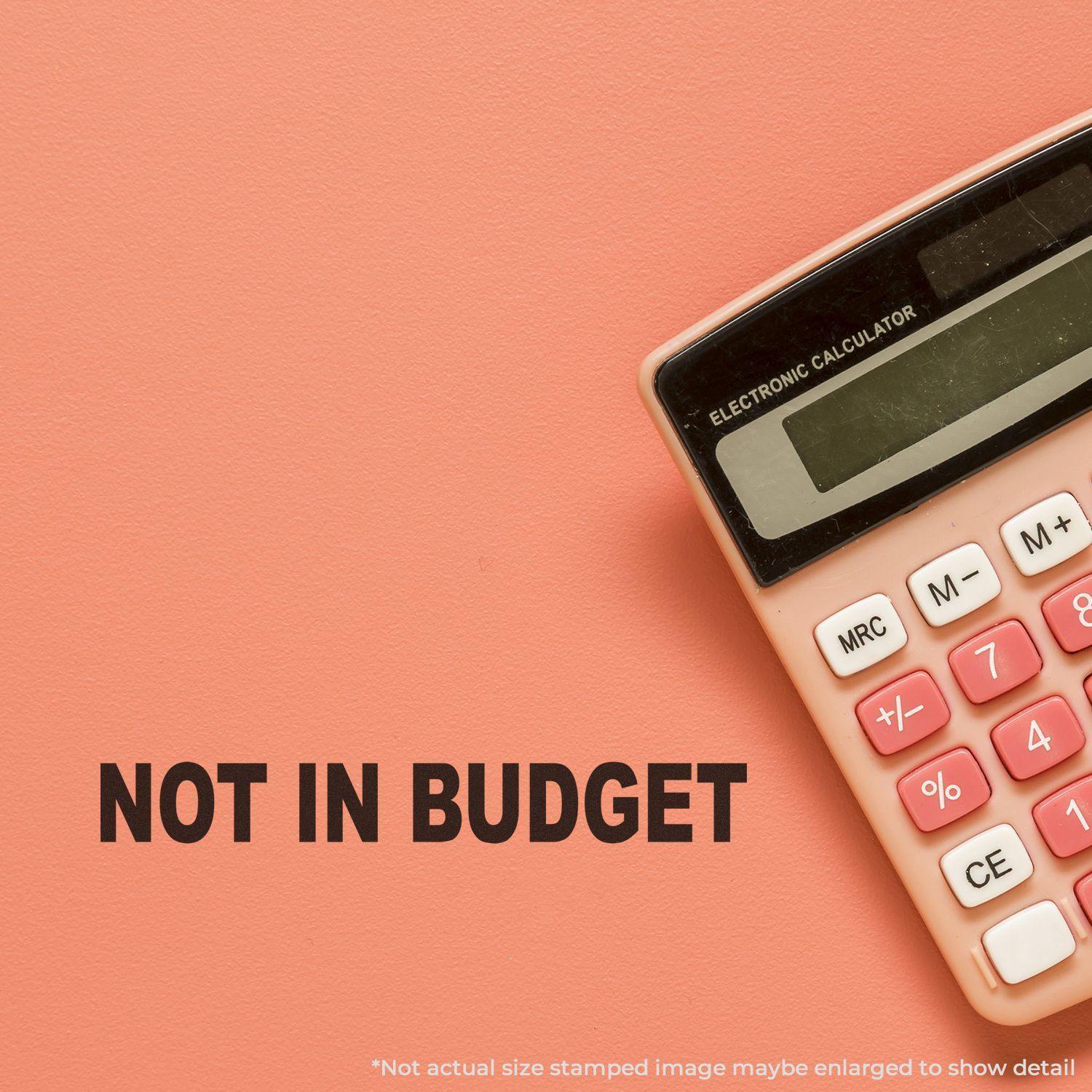 A Large Pre-Inked Not In Budget Stamp is shown on a pink background next to a calculator, with the stamped text NOT IN BUDGET clearly visible.