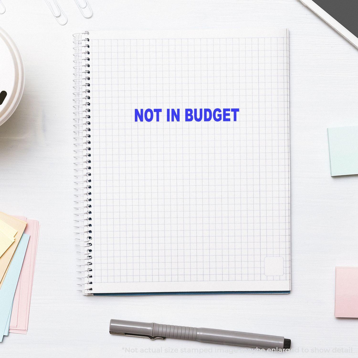 Large Not In Budget Rubber Stamp used on a spiral notebook with grid paper, surrounded by sticky notes, a pen, and paper clips.